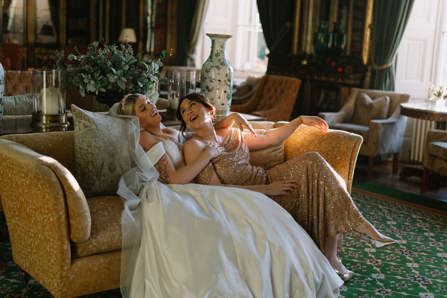 Two brides laughing together on a gold sofa in an elegant room during their wedding day celebration.
