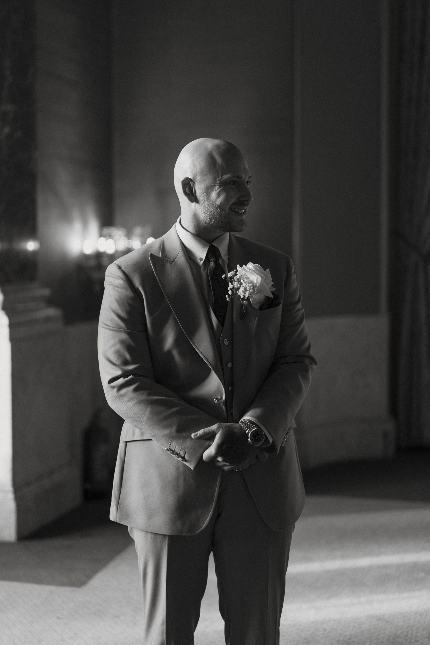 Black and white photo of a bald groom in a suit with boutonniere, smiling at a wedding ceremony.