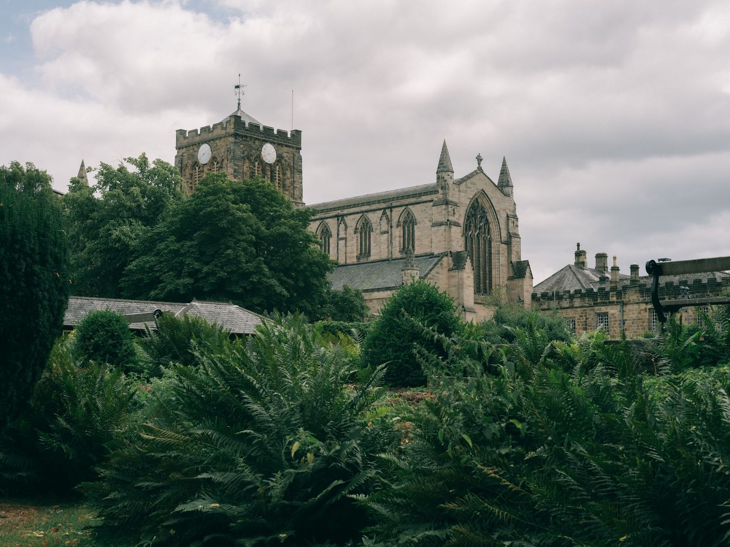 Historic stone cathedral surrounded by lush green trees and ferns under a cloudy sky in England.