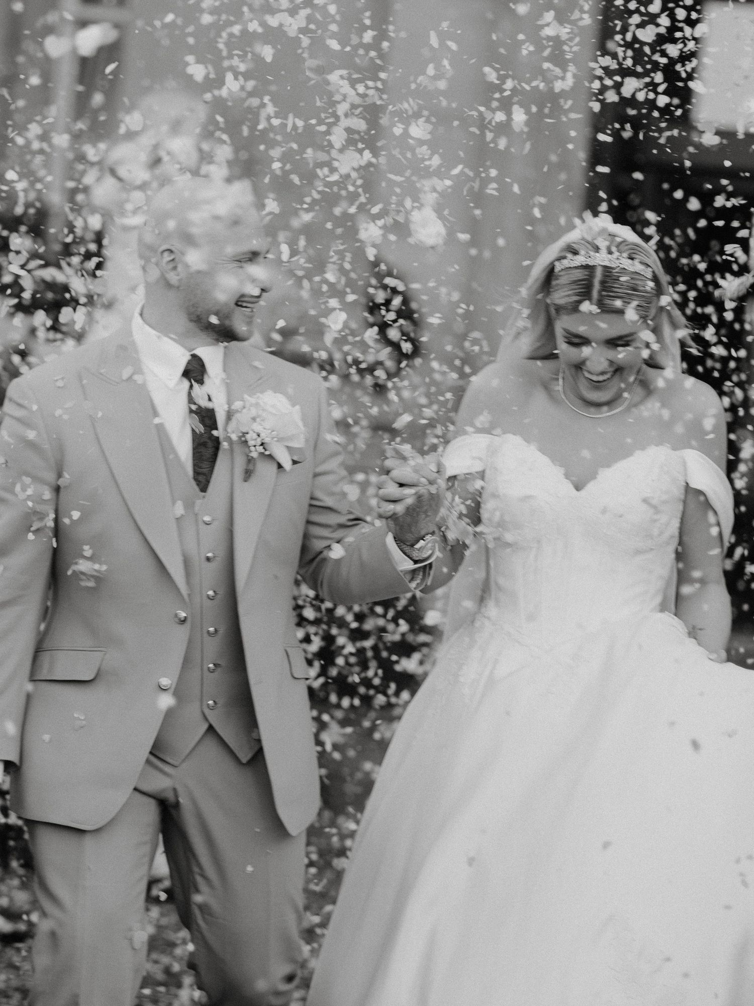 Black and white photo of a smiling bride and groom walking through confetti on their wedding day.