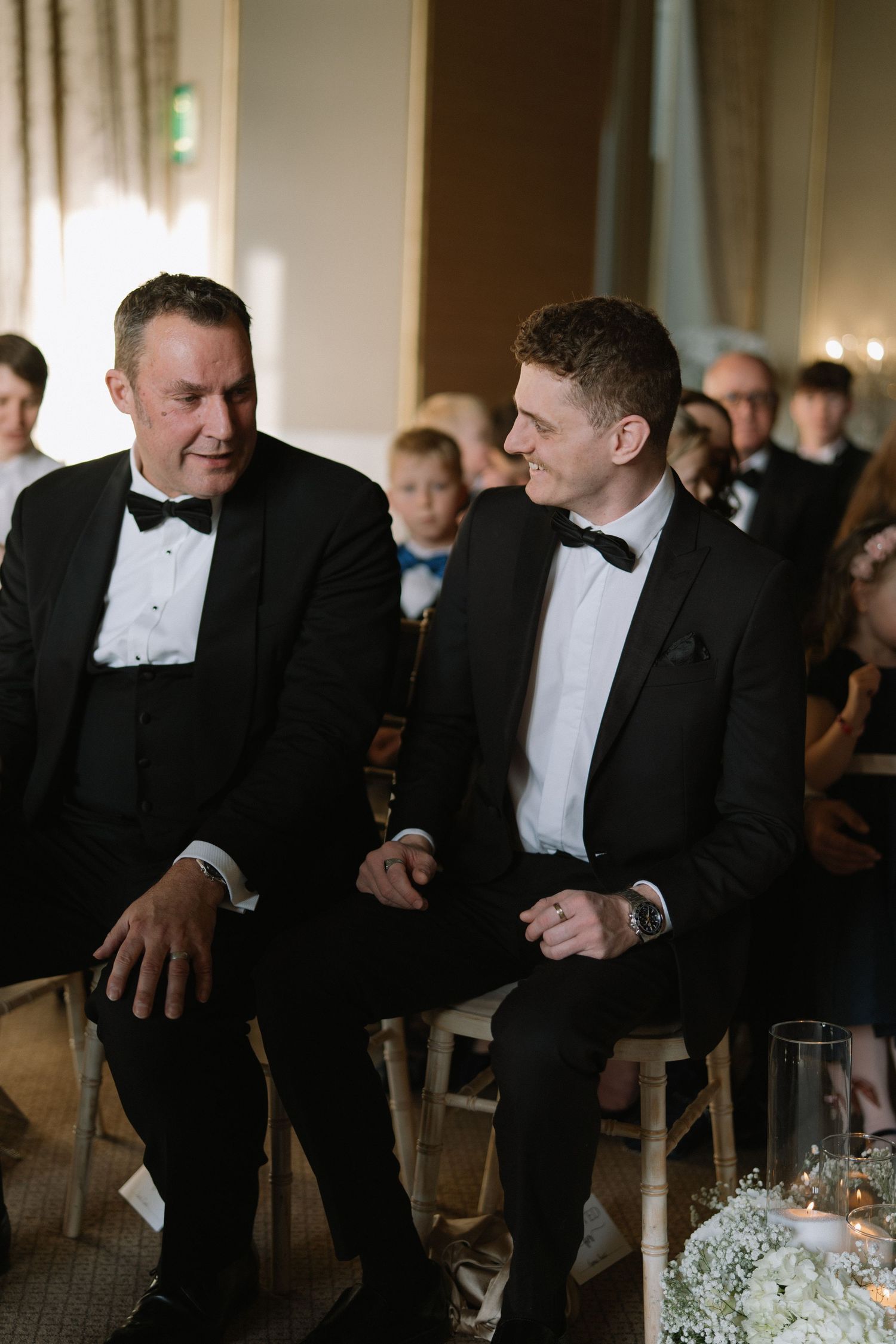 Two men in black tuxedos with bow ties share a laugh while seated at a formal wedding ceremony.
