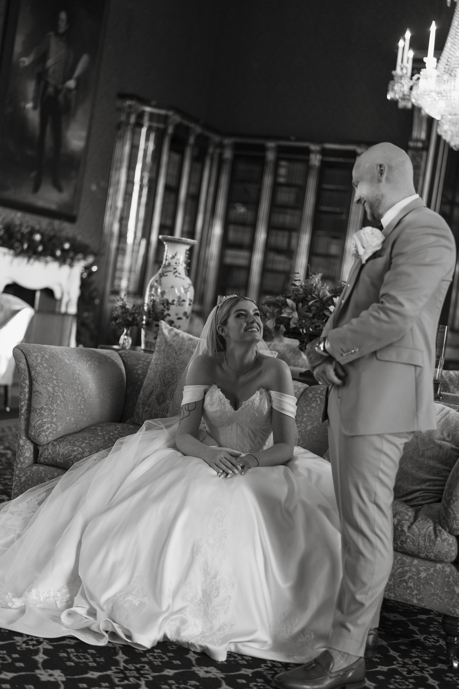 Black and white wedding photo of bride in gown seated on sofa smiling up at bald groom in suit in elegant library.