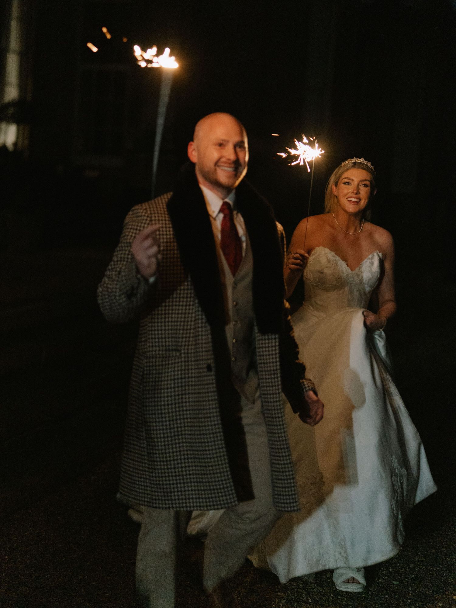 Bride and groom walking at night holding sparklers, bride in white gown, groom in houndstooth coat.
