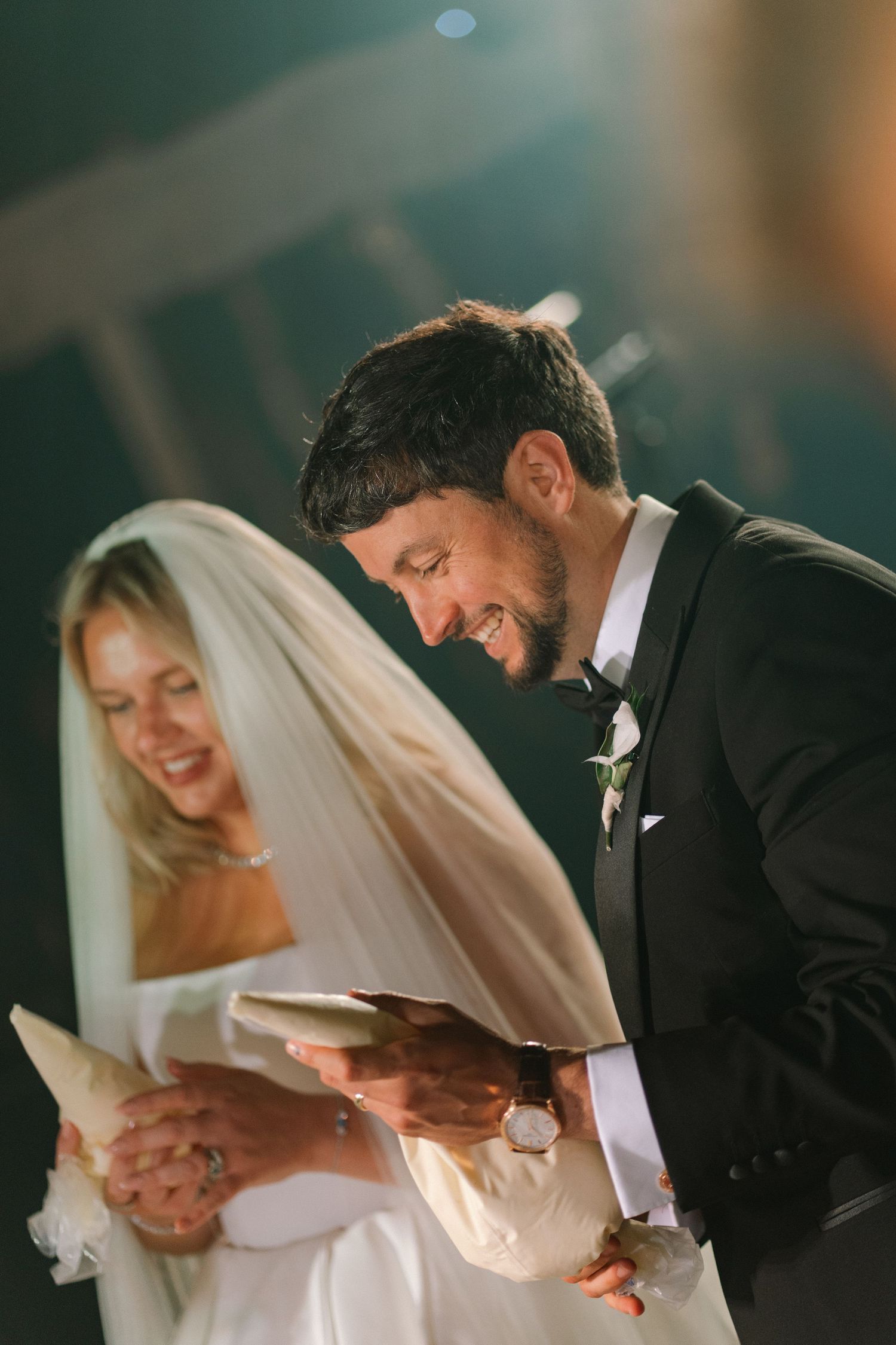 Smiling groom in black tuxedo reads vows beside bride in white wedding dress and veil during ceremony.