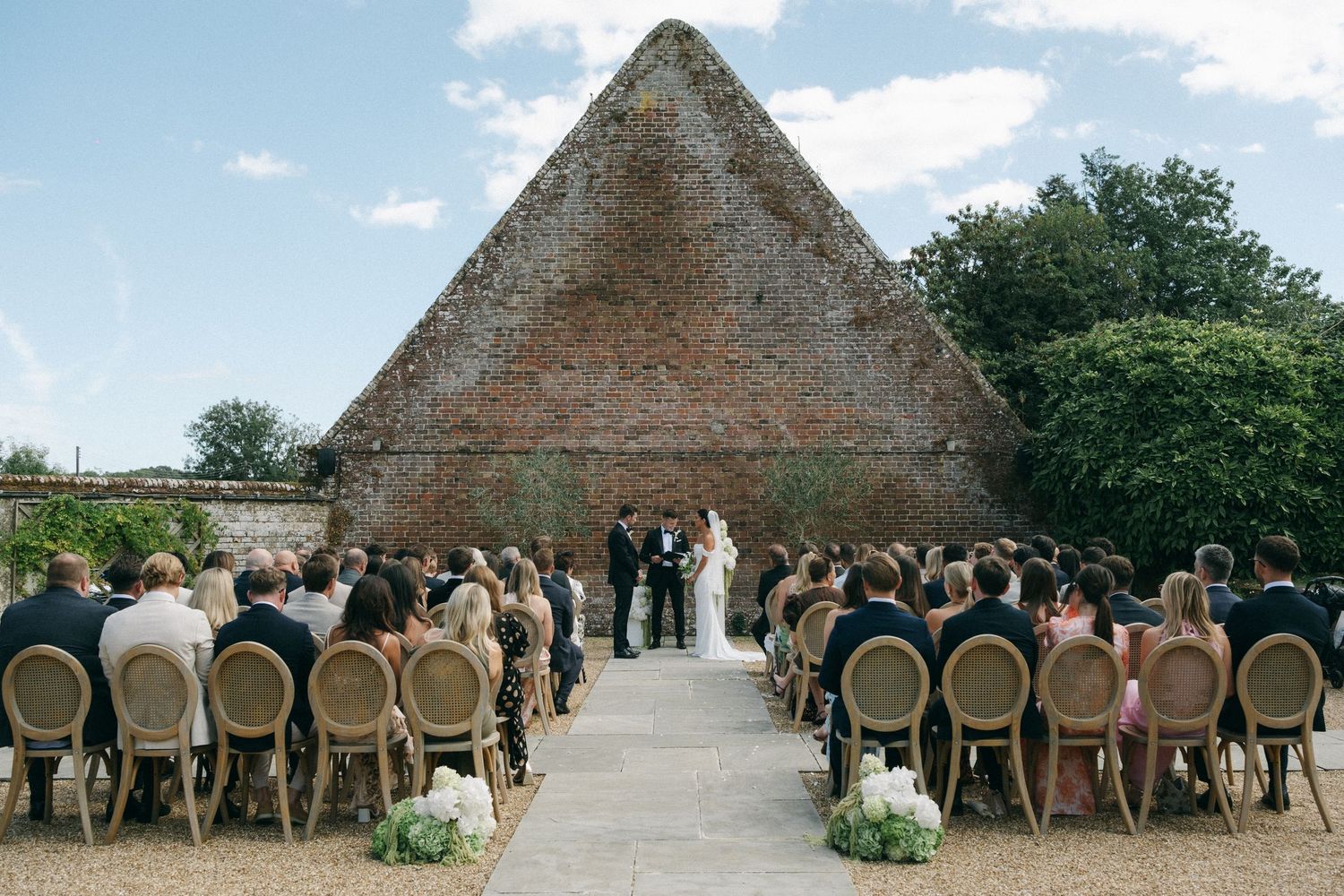 Outdoor wedding ceremony with guests seated in rattan chairs facing couple at altar, brick pyramid structure backdrop.