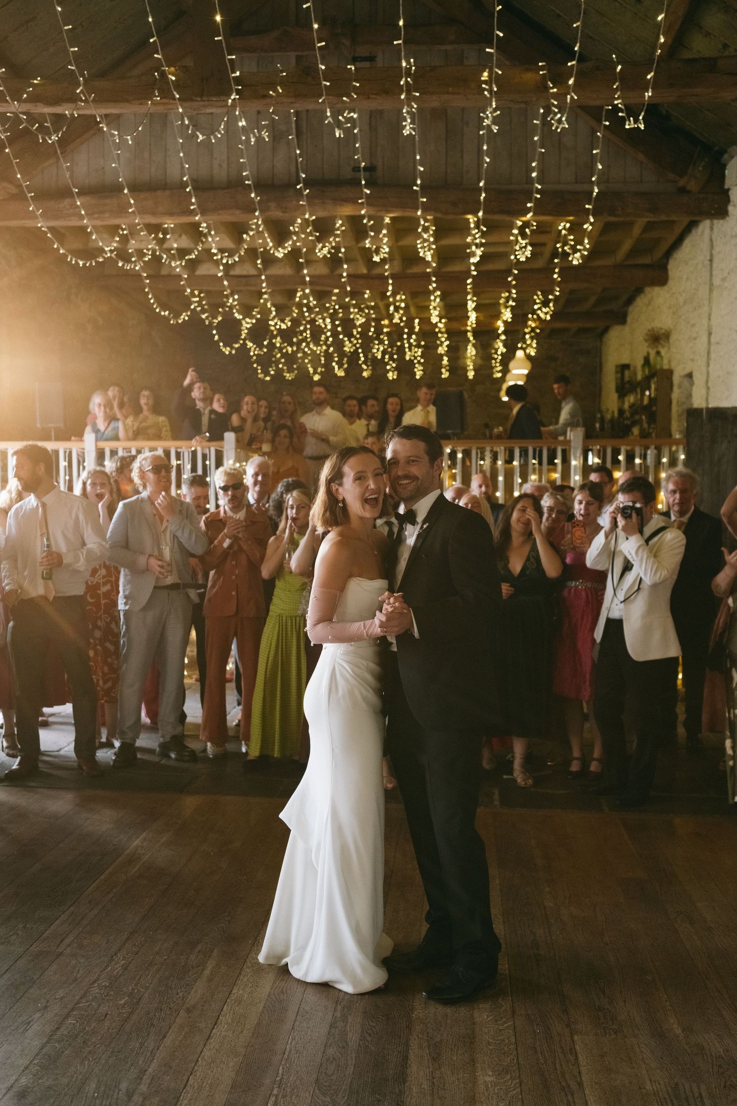 Bride and groom share first dance under fairy lights in rustic barn venue surrounded by applauding wedding guests.