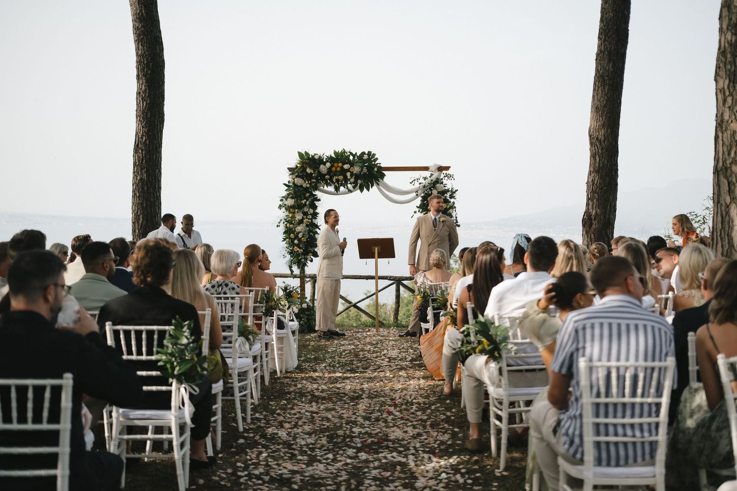Outdoor wedding ceremony with guests seated in white chairs along a petal-lined aisle leading to a floral arch by the sea.