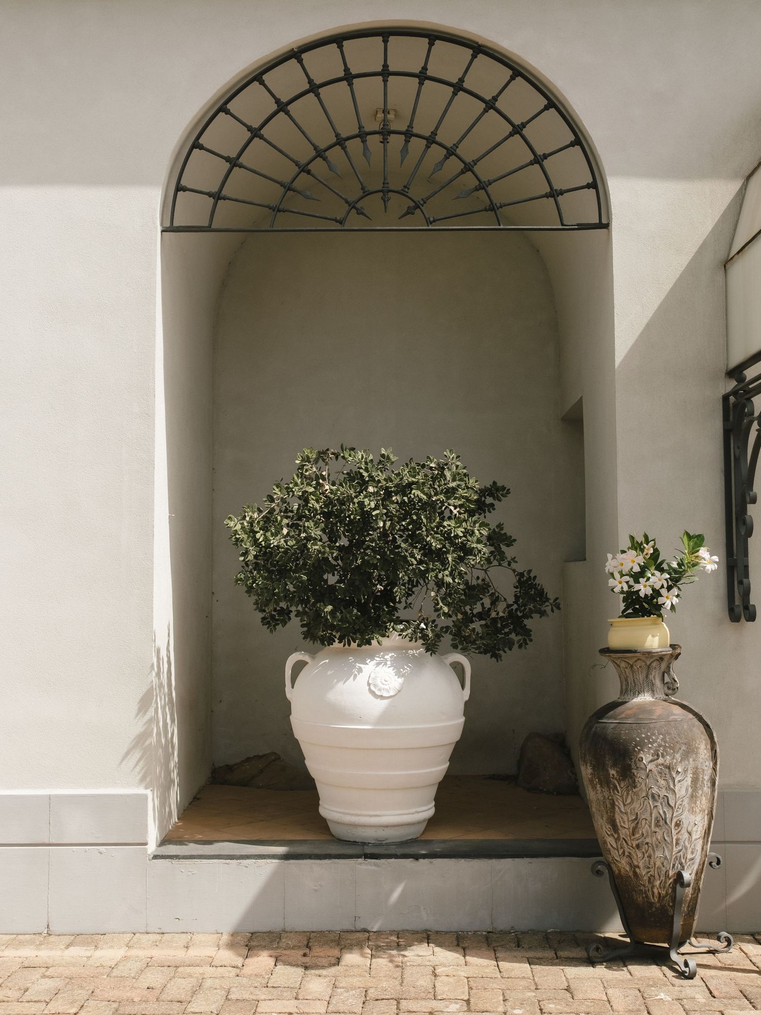 Large white ceramic urn with lush green plant in arched wall niche beside tall bronze vase on brick patio.