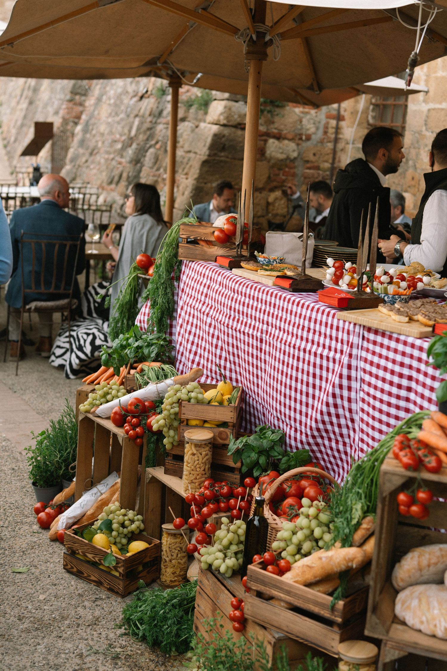 Rustic Italian wedding buffet display with fresh vegetables, fruits, bread, and herbs arranged in wooden crates outdoors.