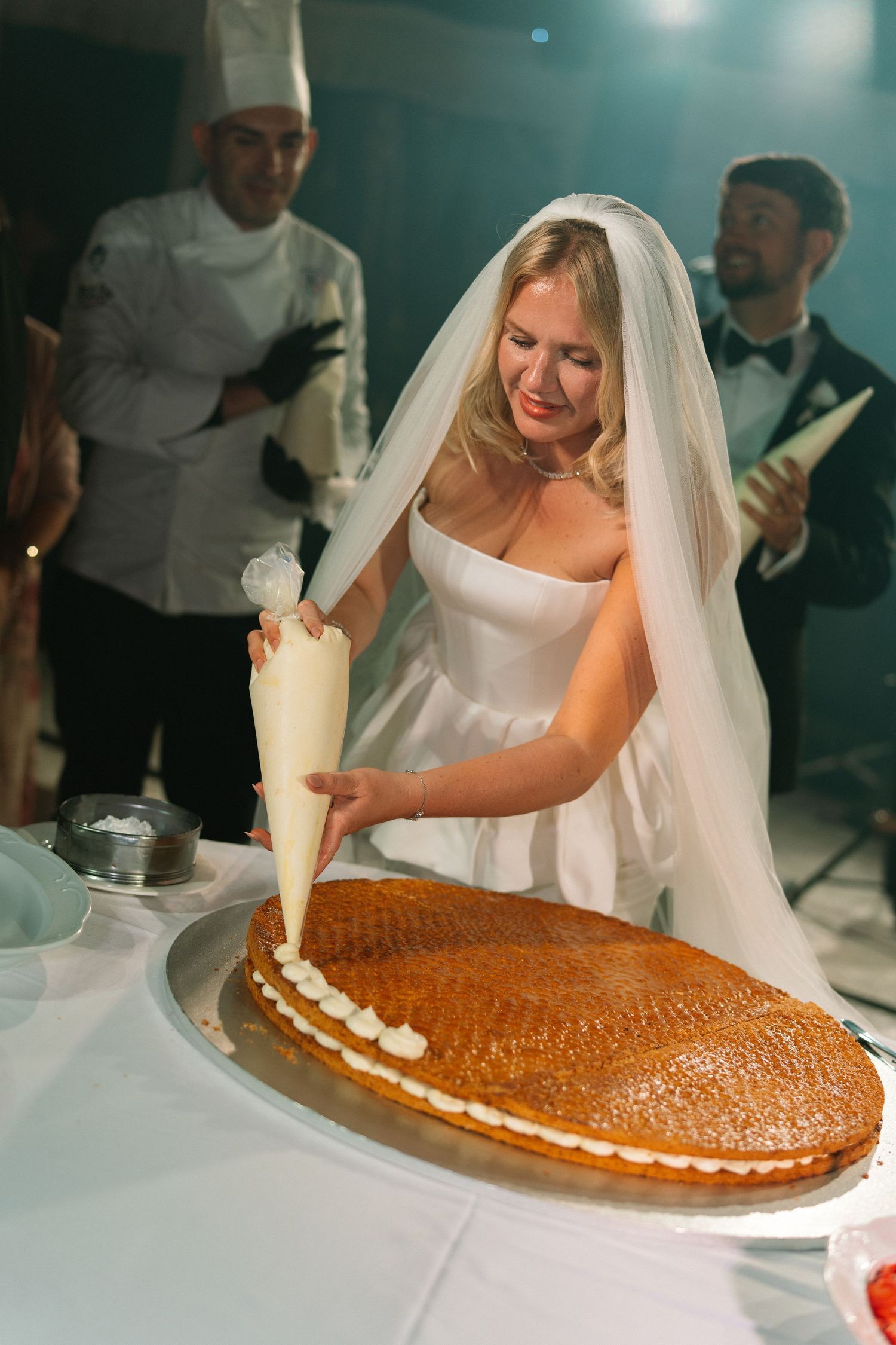Smiling bride in white gown decorating a large wedding cake with cream piping bag.