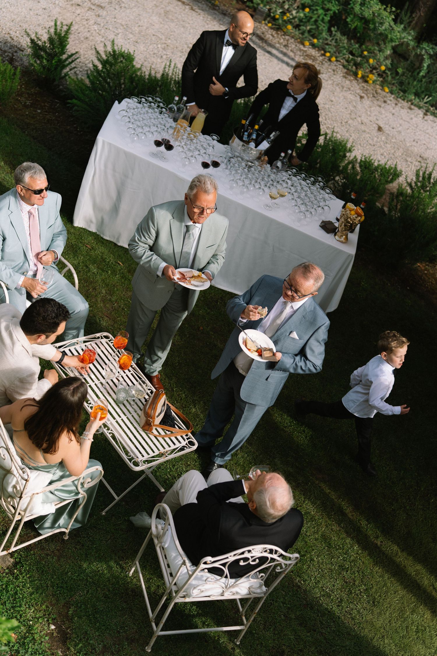 Wedding guests mingle and dine at an elegant outdoor garden reception with white iron furniture.