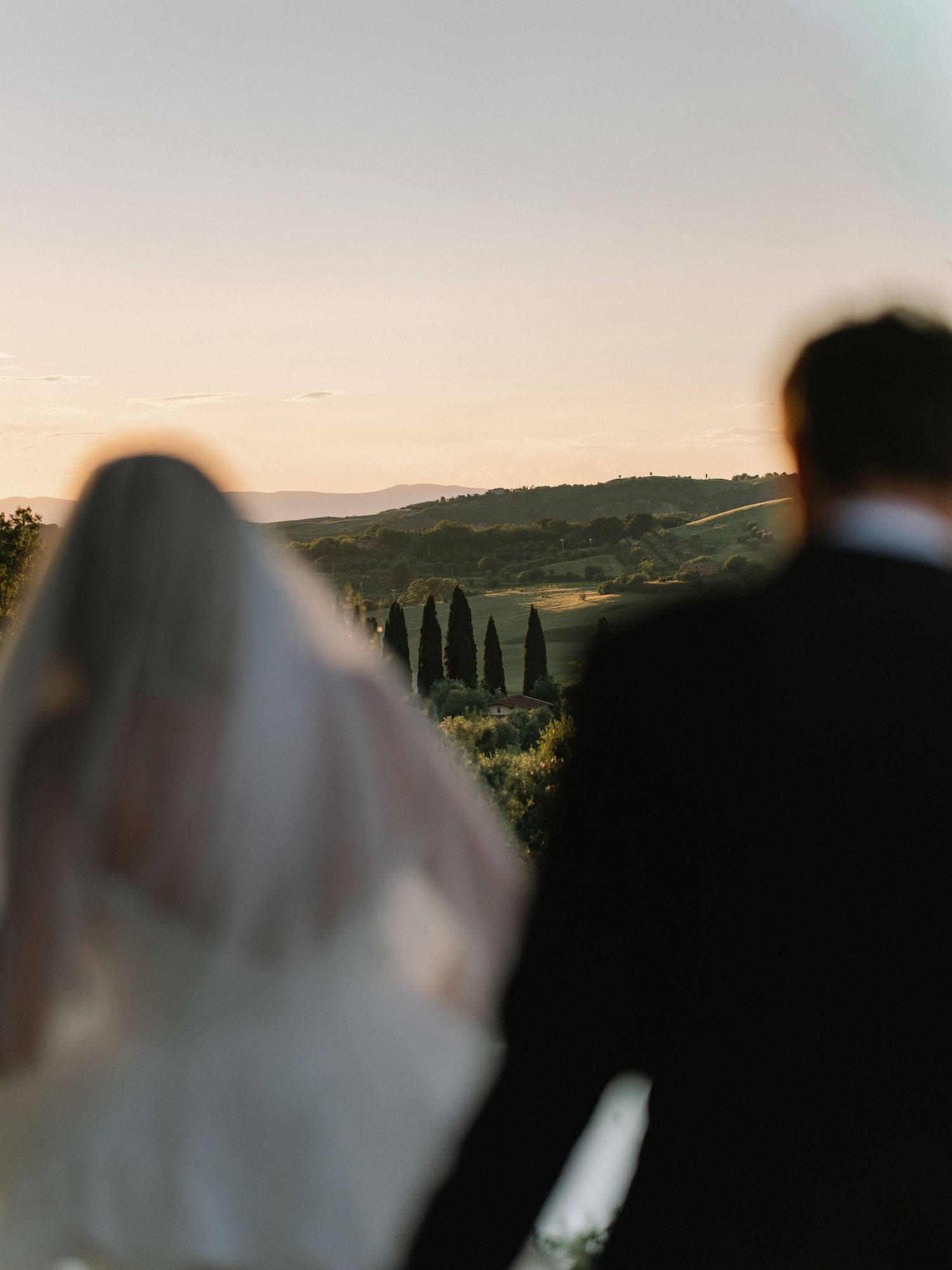 Bride and groom silhouetted against a golden sunset over rolling Tuscan hills with cypress trees.