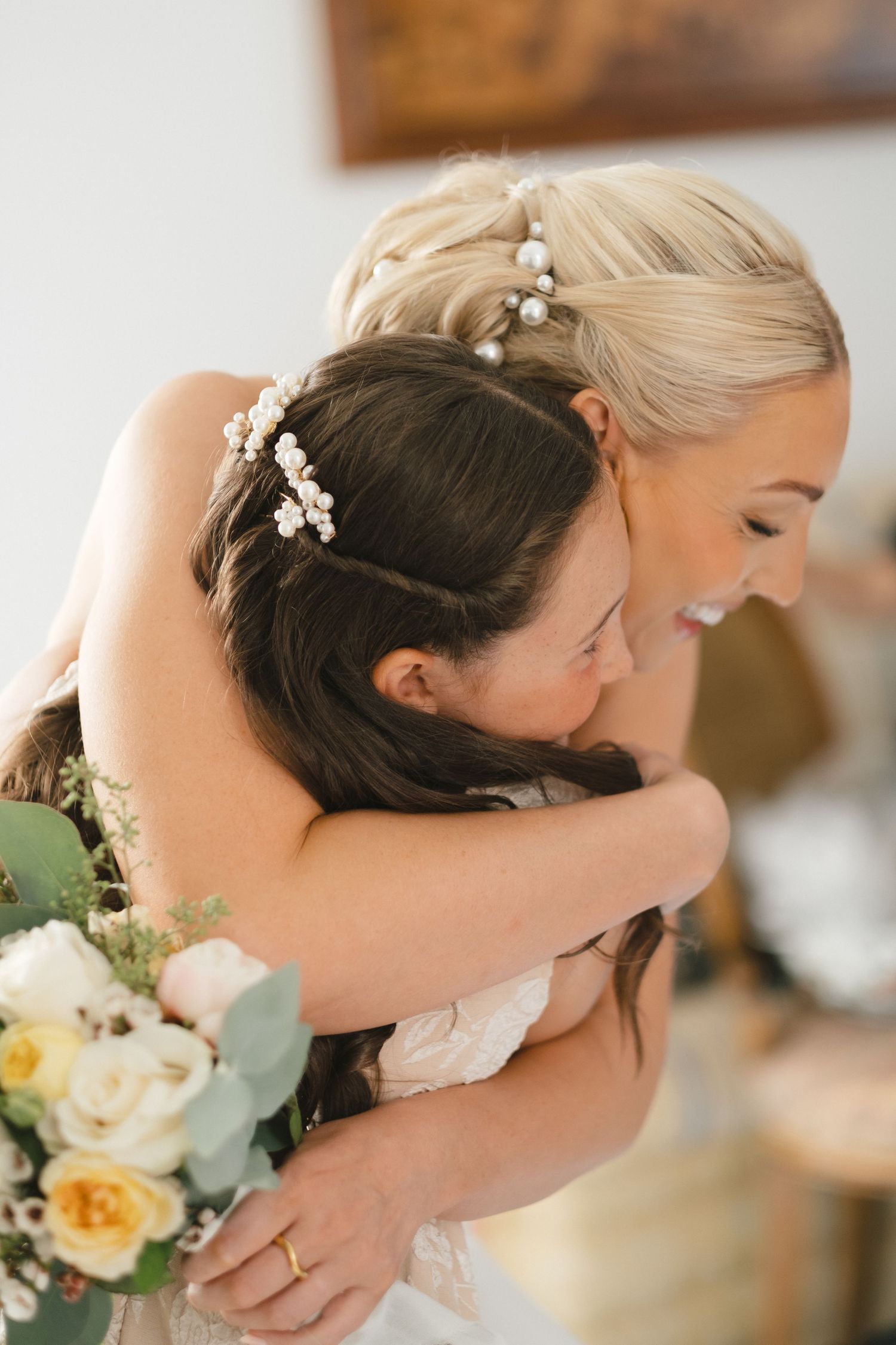 Two brides embracing and laughing joyfully, both wearing floral hair accessories, one holding a bouquet of white and yellow roses.