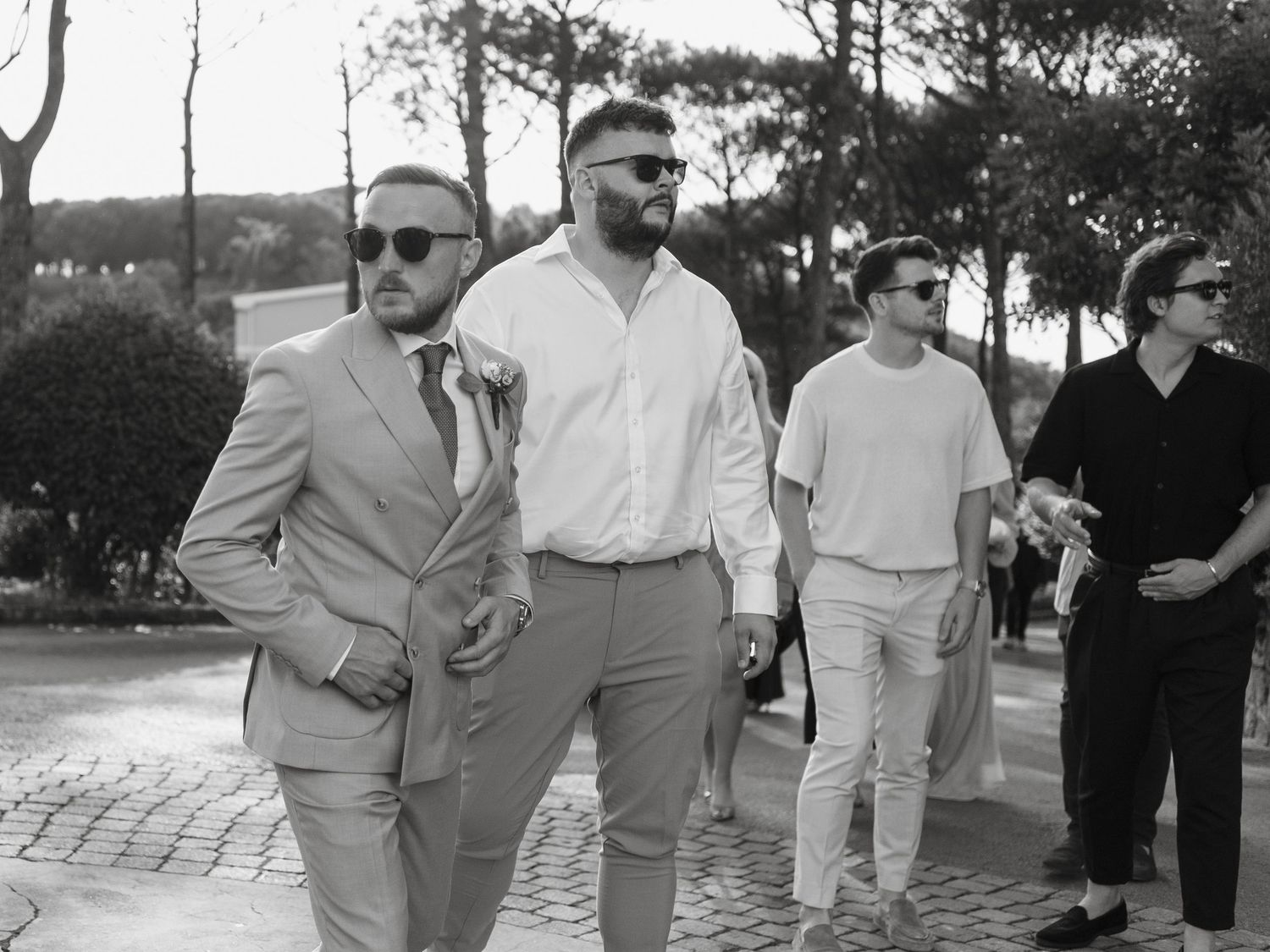Black and white photo of well-dressed men in suits and sunglasses walking along a brick path outdoors.