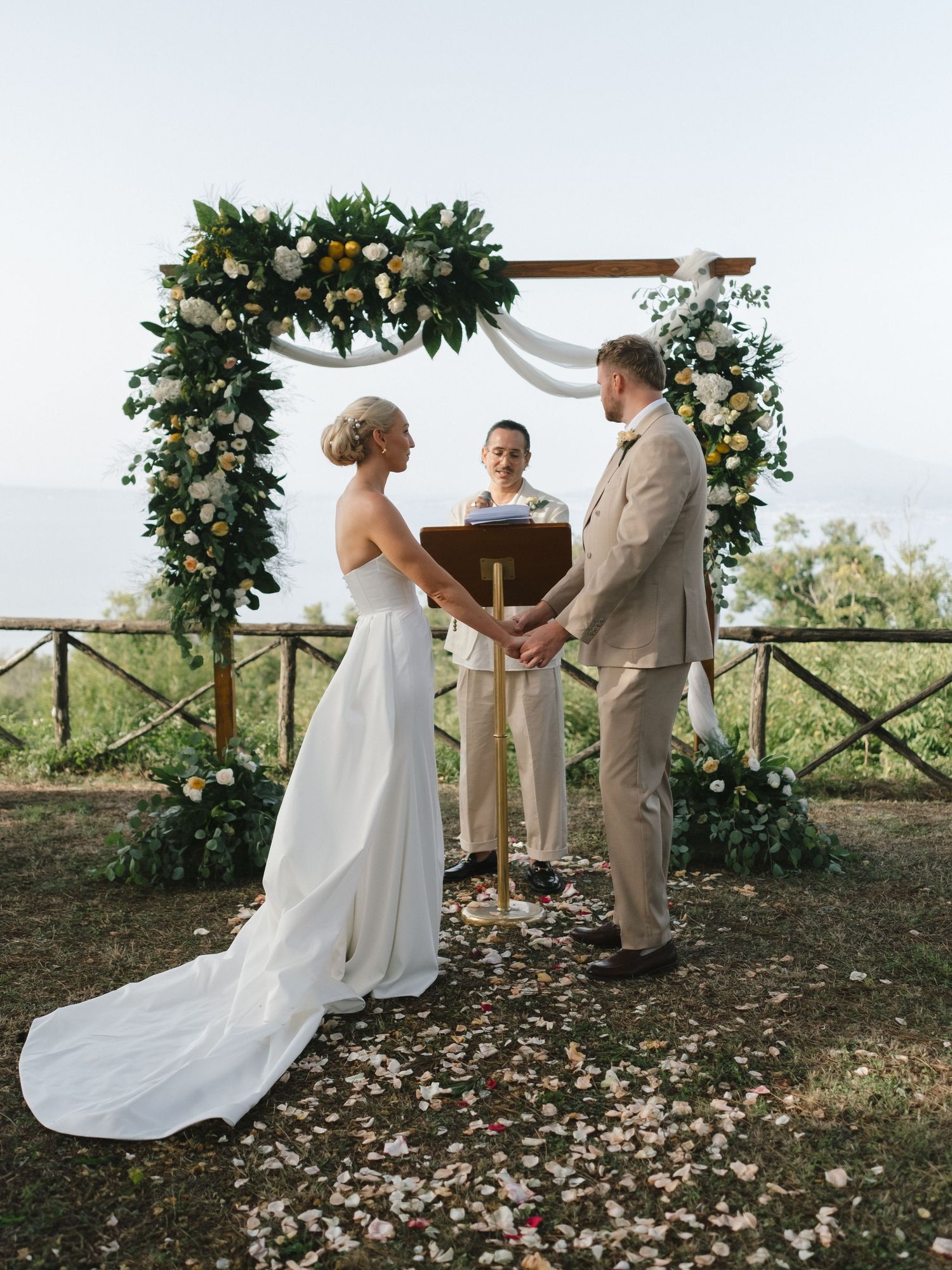 Bride and groom exchange vows under floral arch with ocean view at elegant outdoor wedding ceremony.