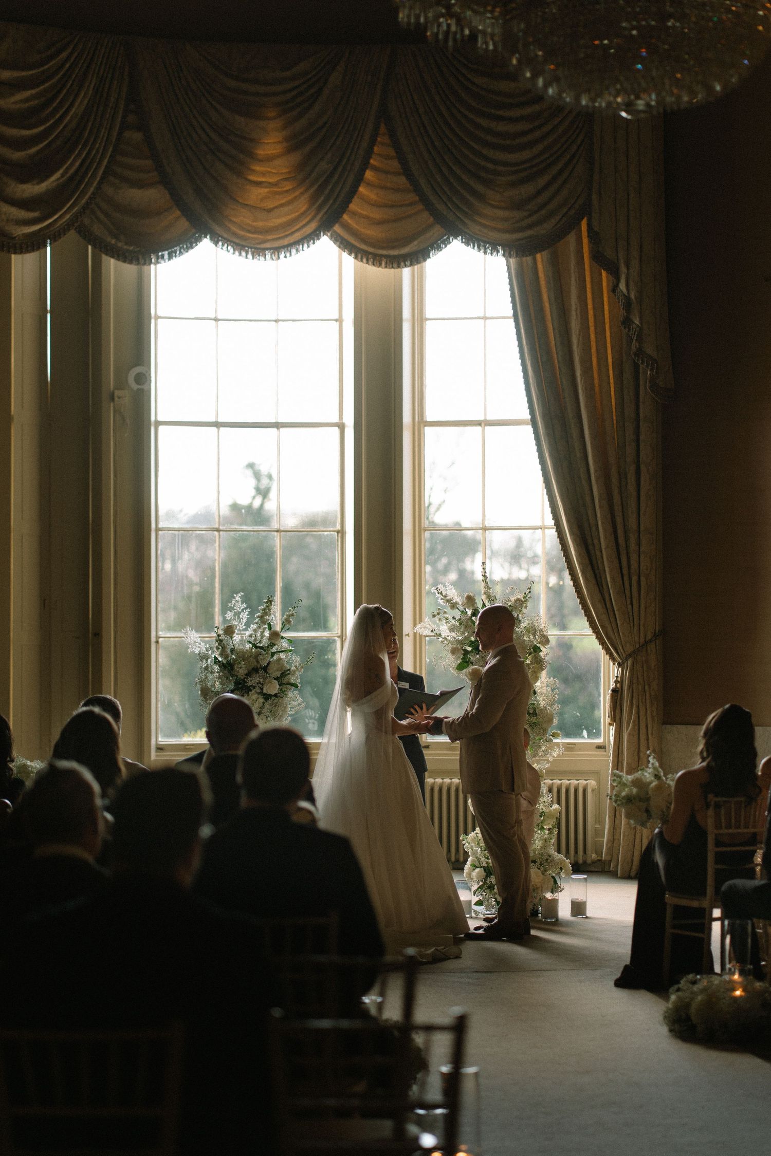Elegant wedding ceremony in a grand room with tall windows, couple exchanging vows before floral arch and seated guests.