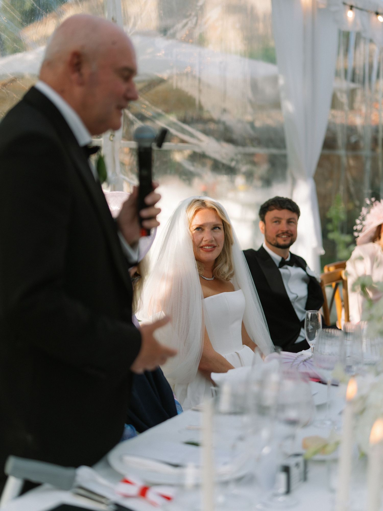 Bride in white gown and veil smiles at wedding reception as speaker gives toast at elegant candlelit table.