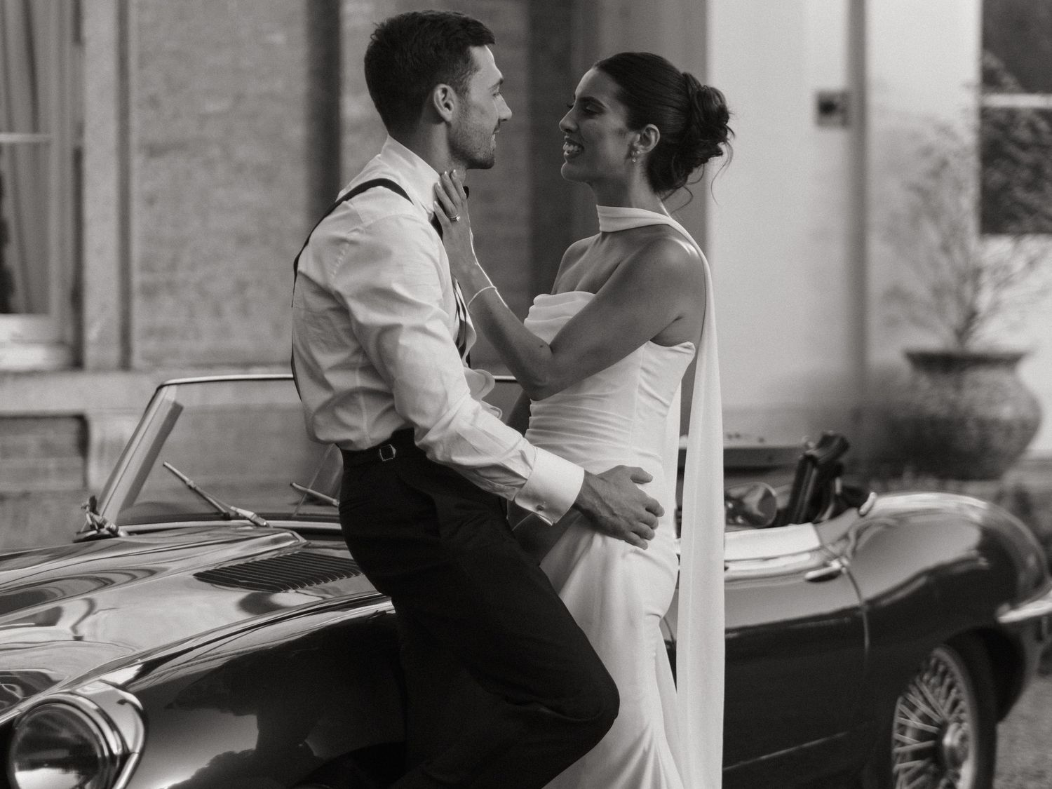 Black and white photo of a bride and groom embracing beside a classic vintage car on their wedding day.