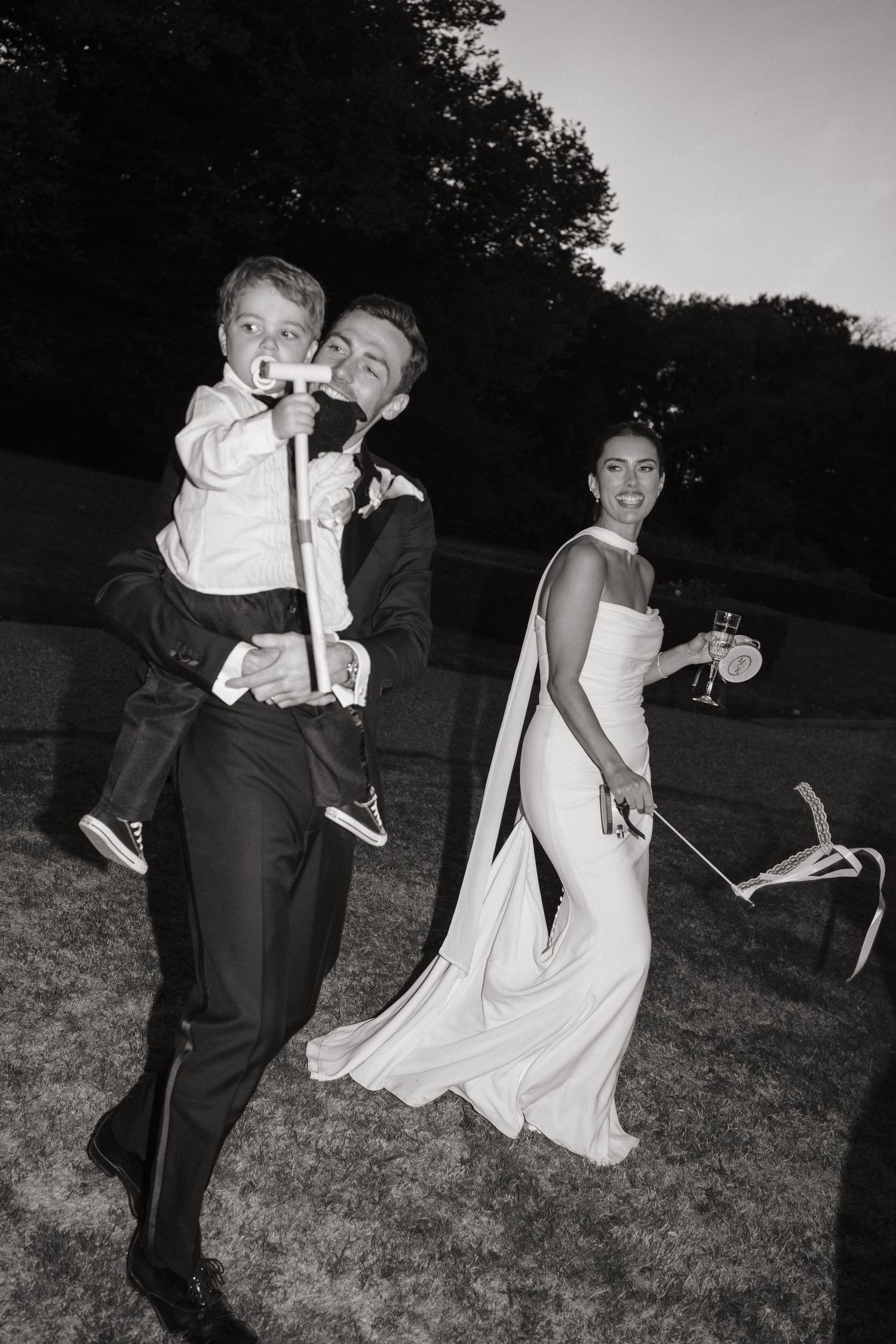 Black and white photo of a groom carrying a young boy while bride in white gown walks beside them at outdoor wedding.