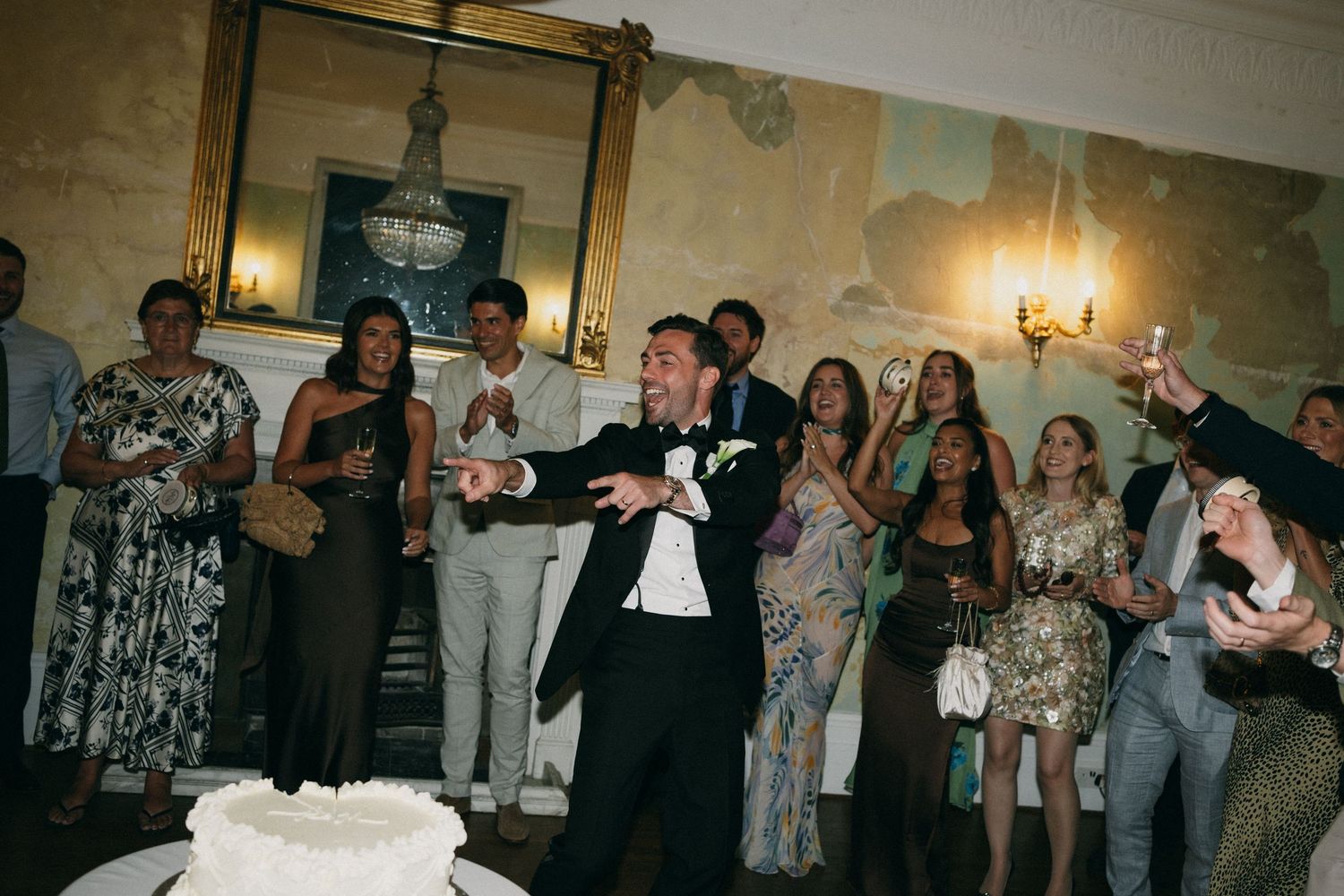 Groom celebrating at wedding reception with guests cheering, white cake visible, elegant venue with chandelier.