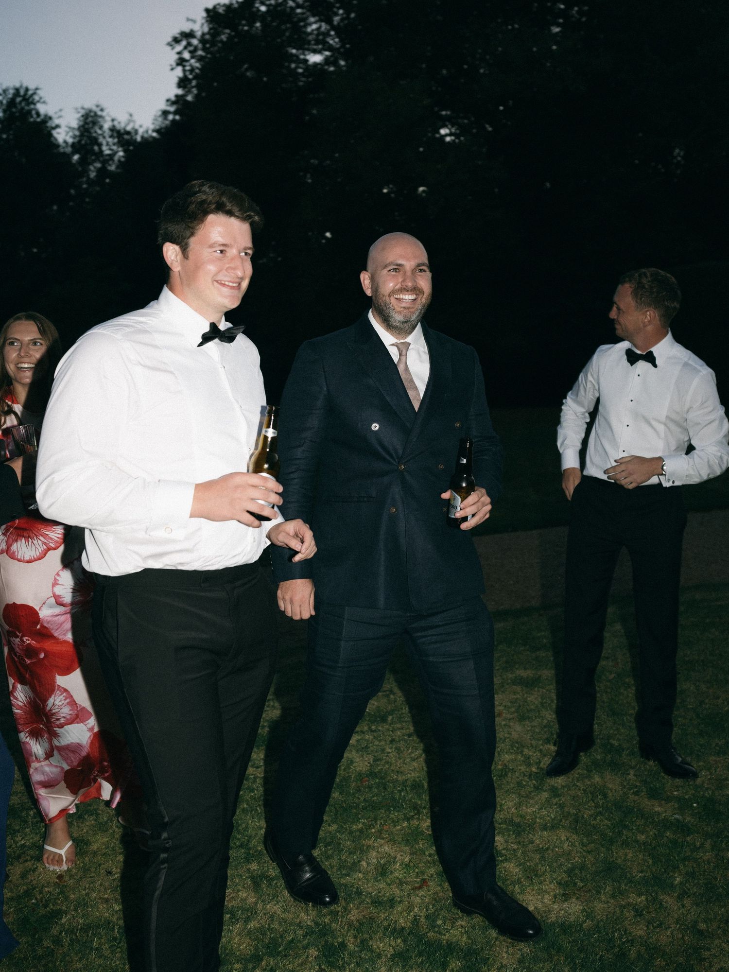 Two men in formal attire laughing and holding drinks at an outdoor evening wedding reception.