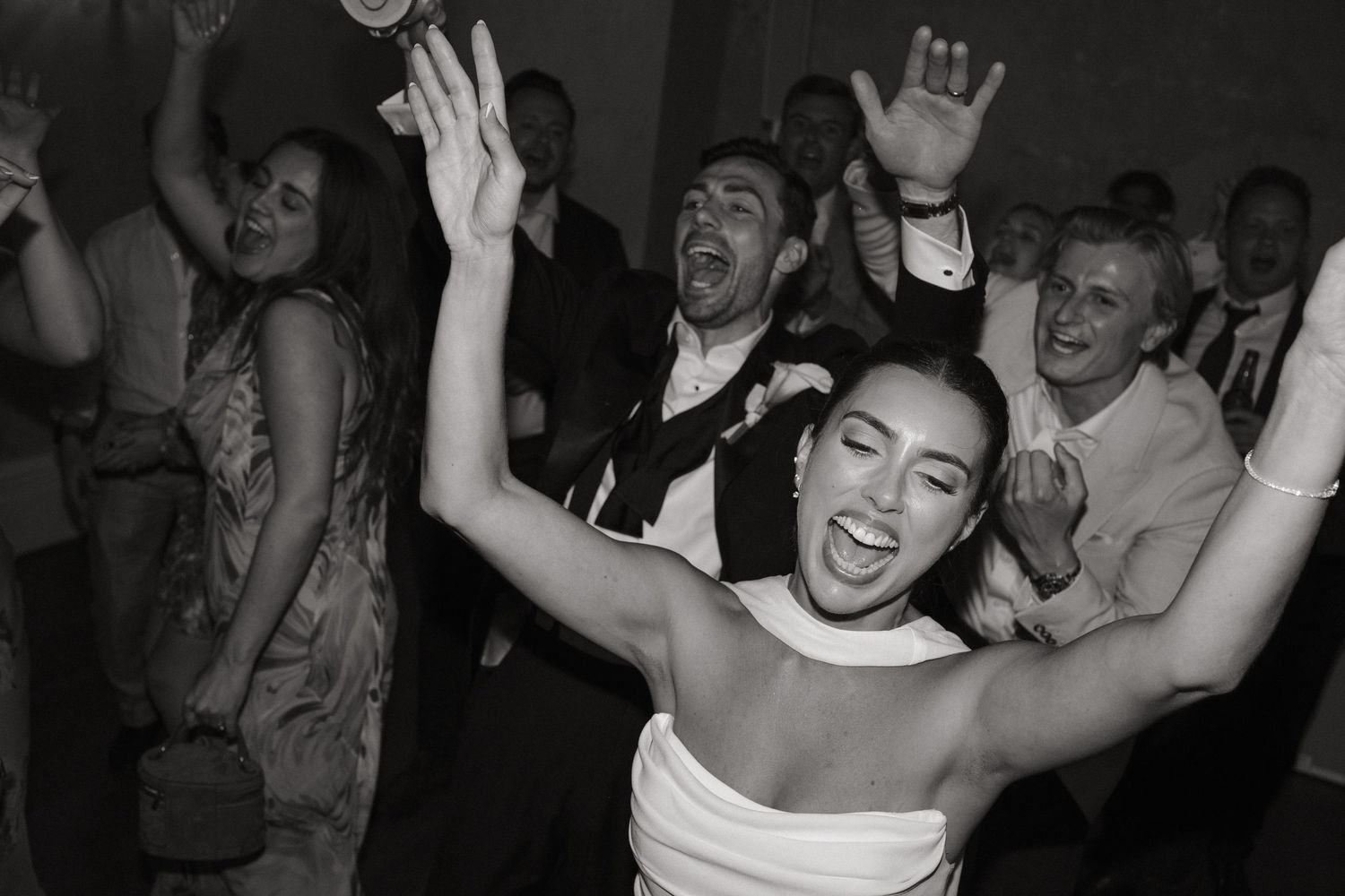 Black and white photo of a joyful bride dancing with hands raised at a lively wedding reception.