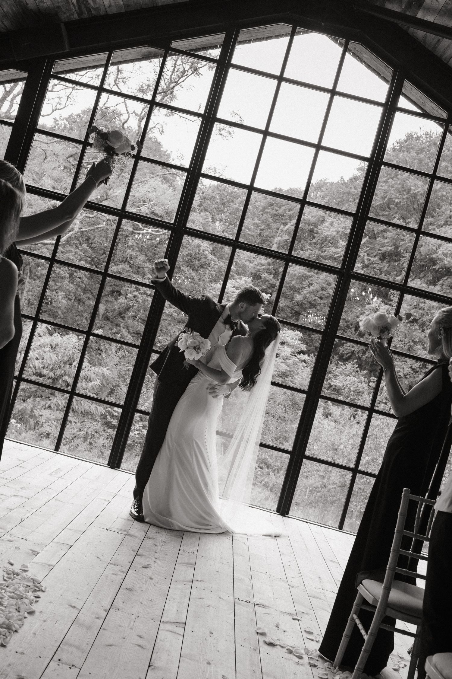 Black and white photo of a bride and groom sharing a kiss at the altar before a grand arched window.