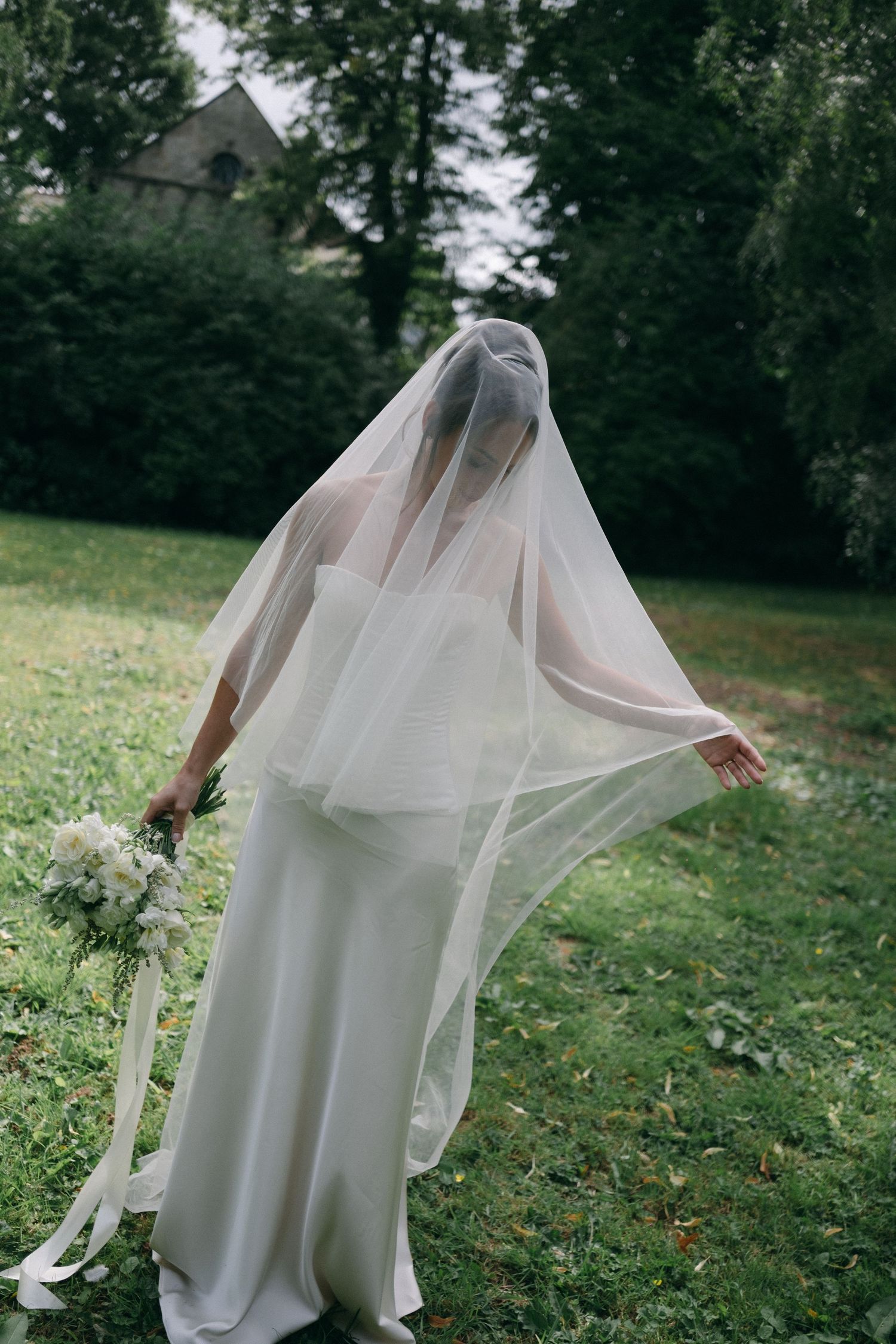 Bride in elegant white gown with flowing veil draped over head, holding bouquet outdoors in lush garden setting.