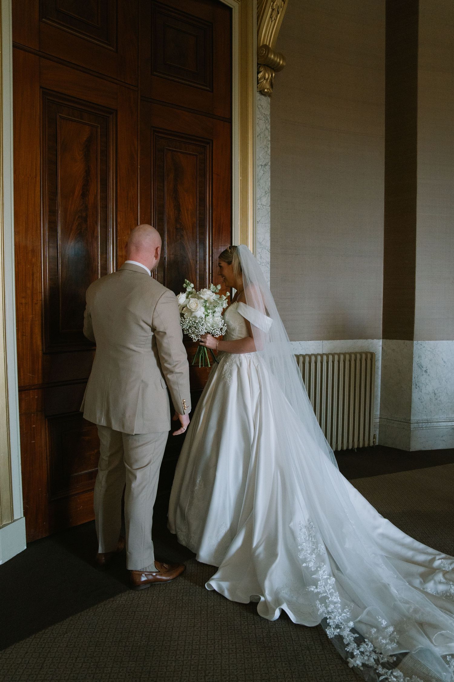 Bride in white gown with lace train and groom in tan suit stand before ornate wooden doors in elegant venue.
