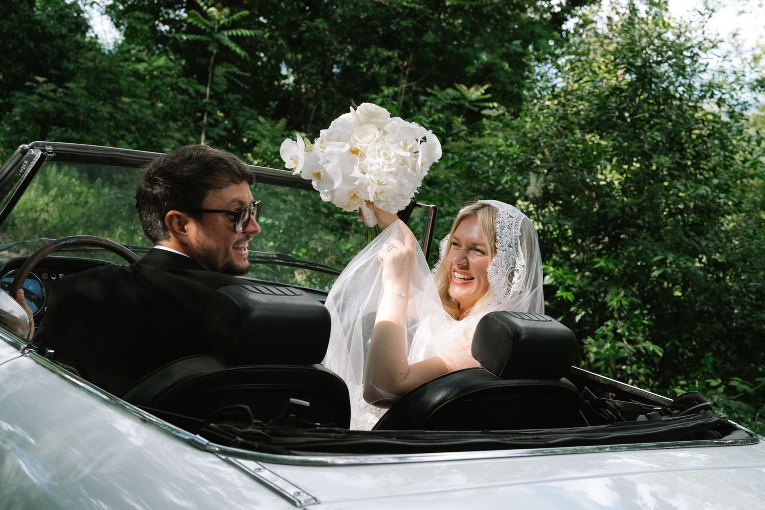 Bride holding white bouquet and groom in sunglasses ride in a vintage white convertible wedding car.