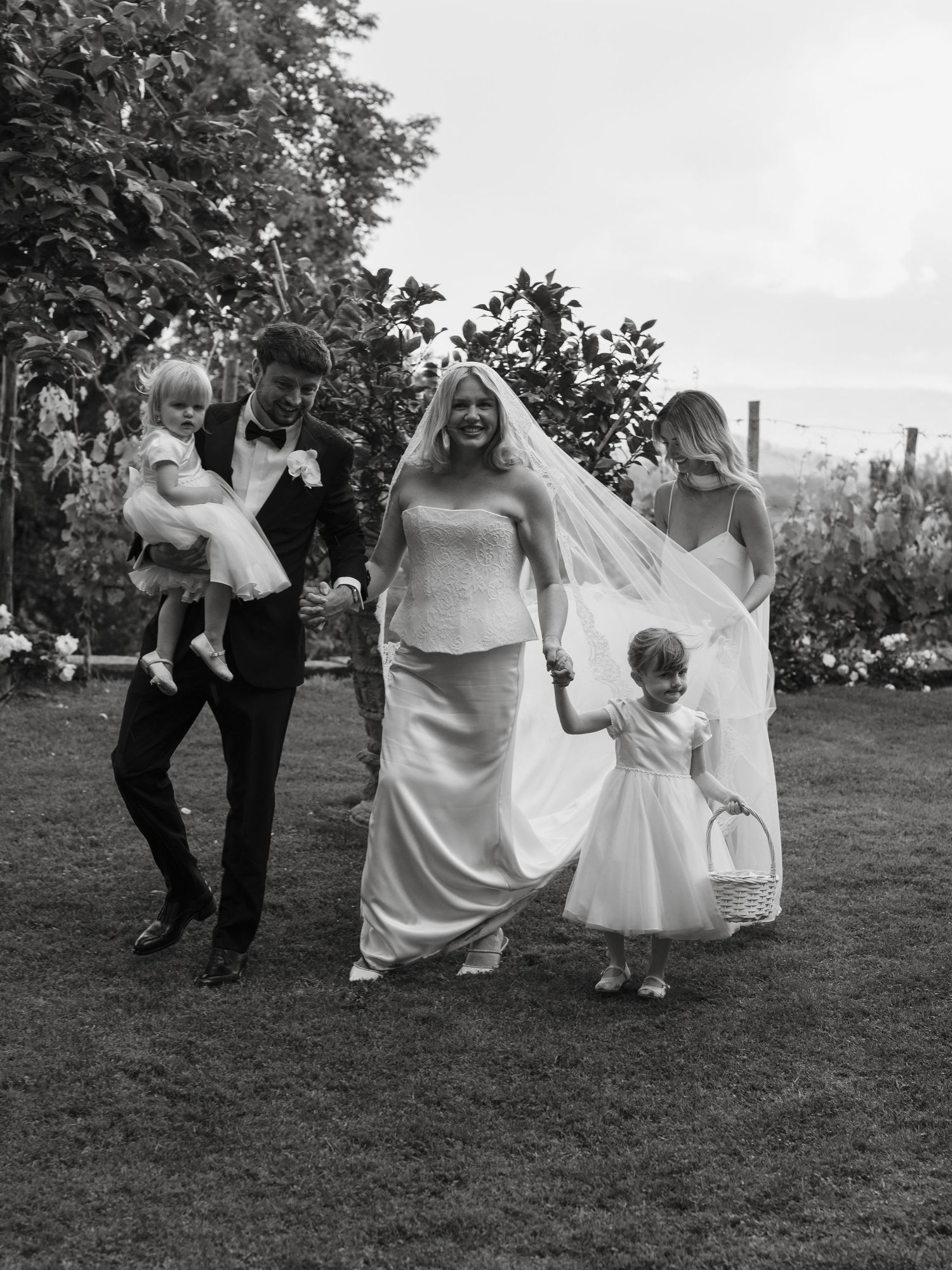 Black and white photo of bride and groom walking with flower girl and child in garden setting.