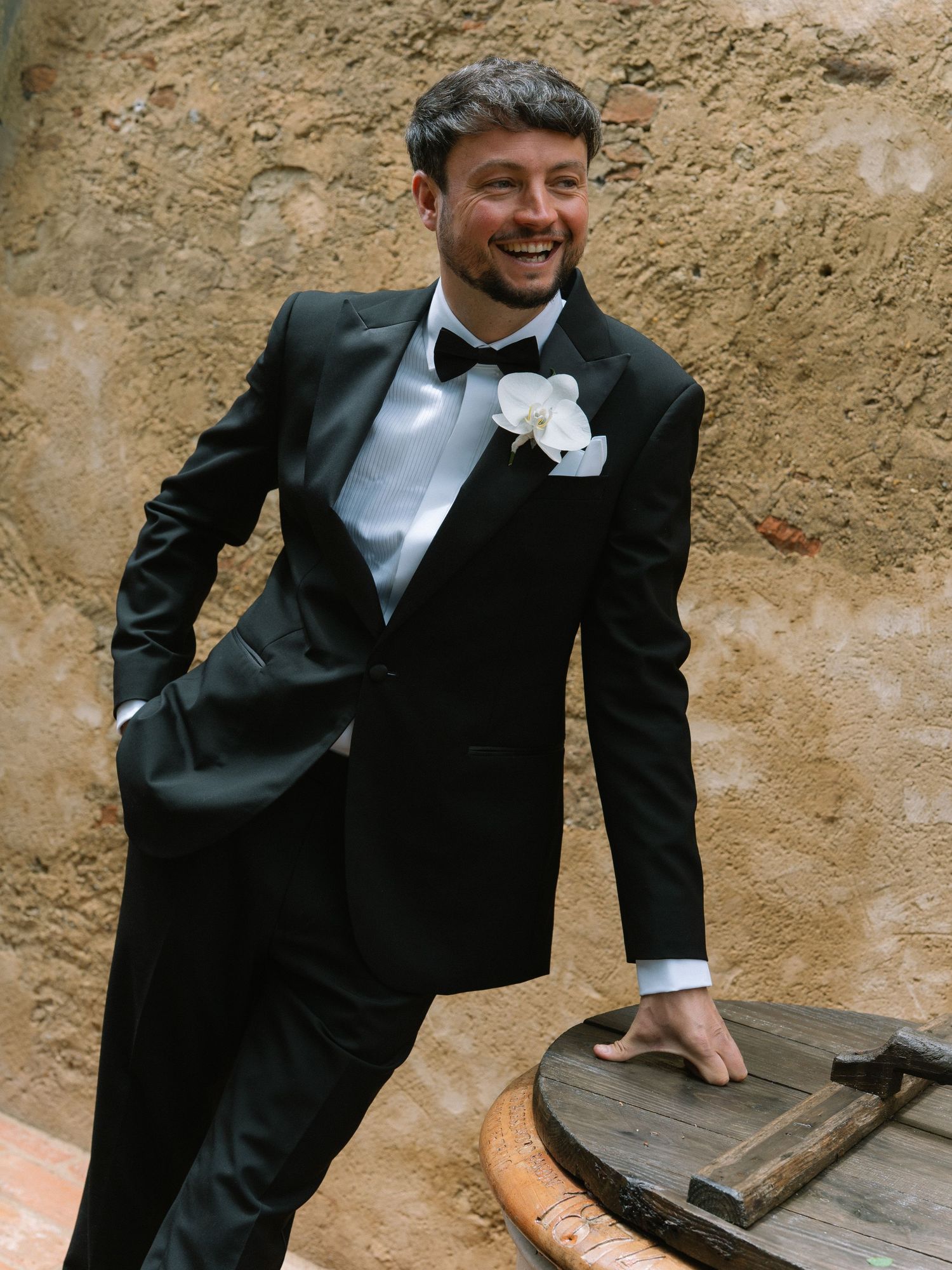 Smiling groom in black tuxedo with white orchid boutonniere leaning against a rustic stone wall.