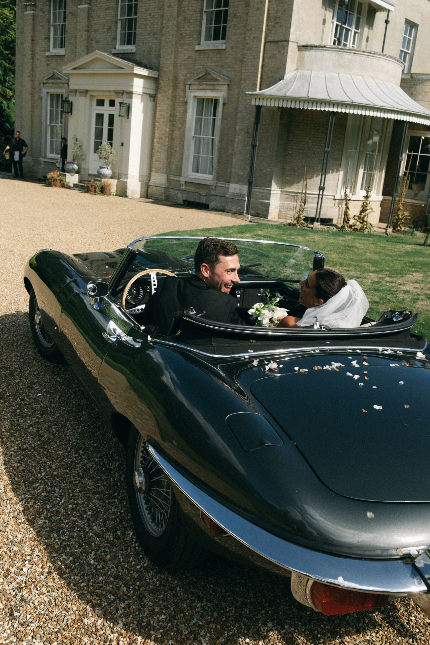 Bride and groom riding in a classic dark green convertible Jaguar E-Type outside an elegant country house venue.