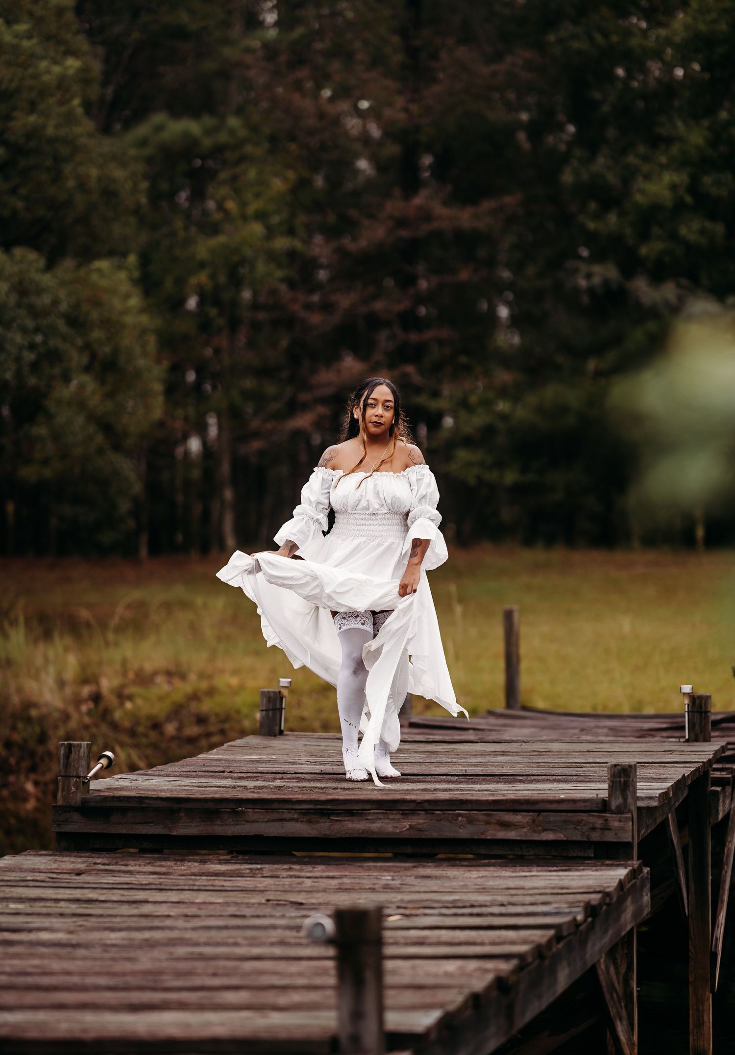 Woman in flowing white off-shoulder dress walking confidently on wooden dock with green forest backdrop.
