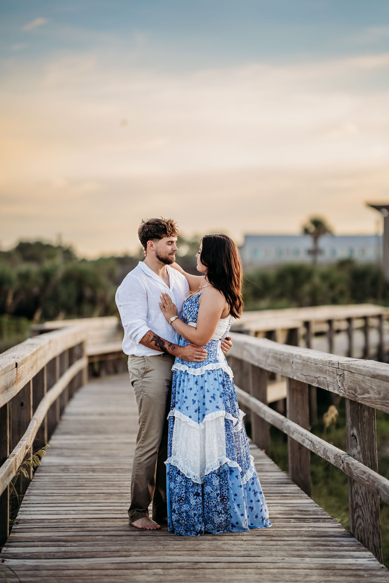 Couple embracing on a wooden boardwalk at sunset, woman in blue floral maxi dress, man in white shirt and khaki pants.