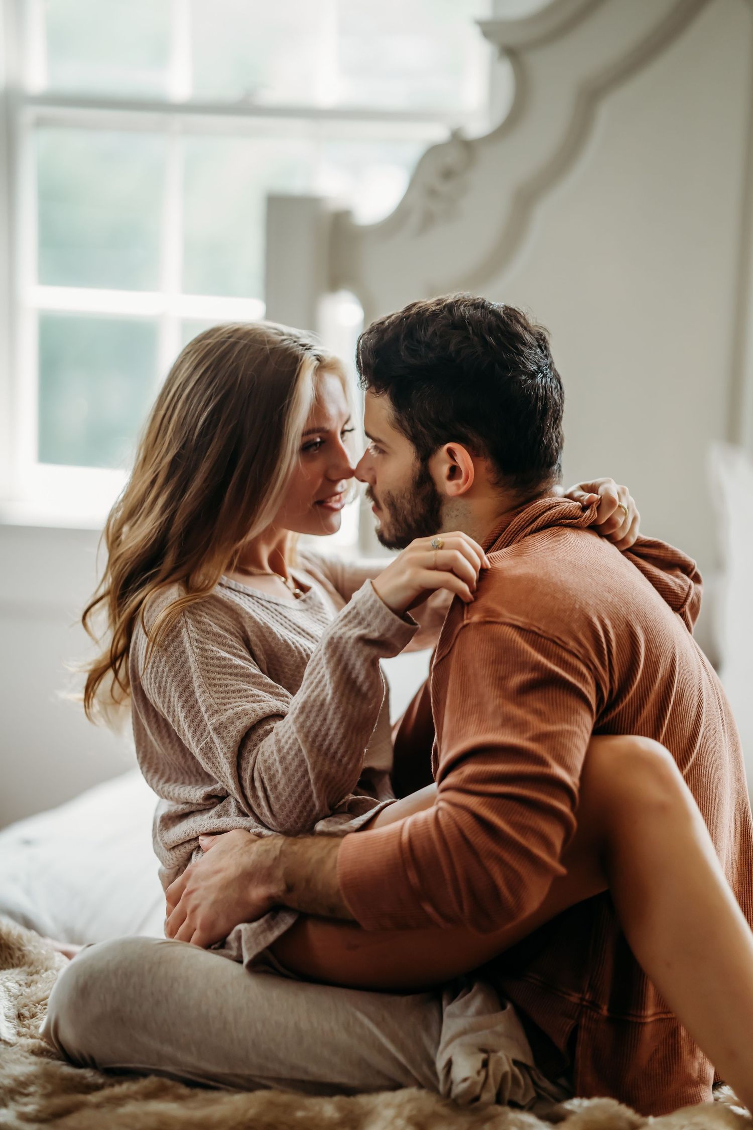 Romantic couple sitting face to face on a bed, smiling and holding hands intimately.
