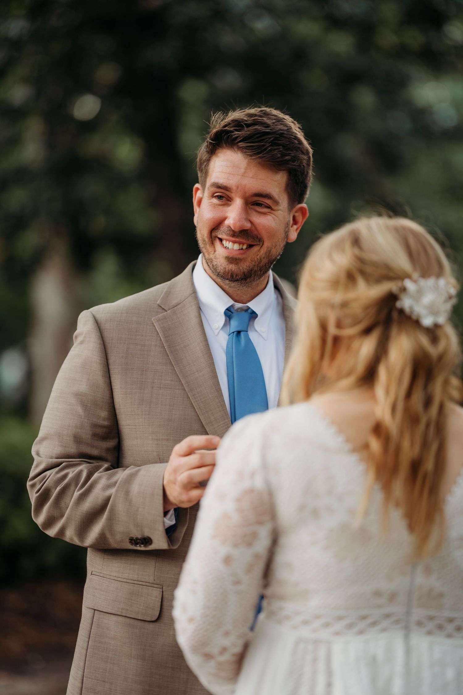 Smiling groom in tan suit and blue tie looks lovingly at blonde bride during outdoor wedding ceremony.