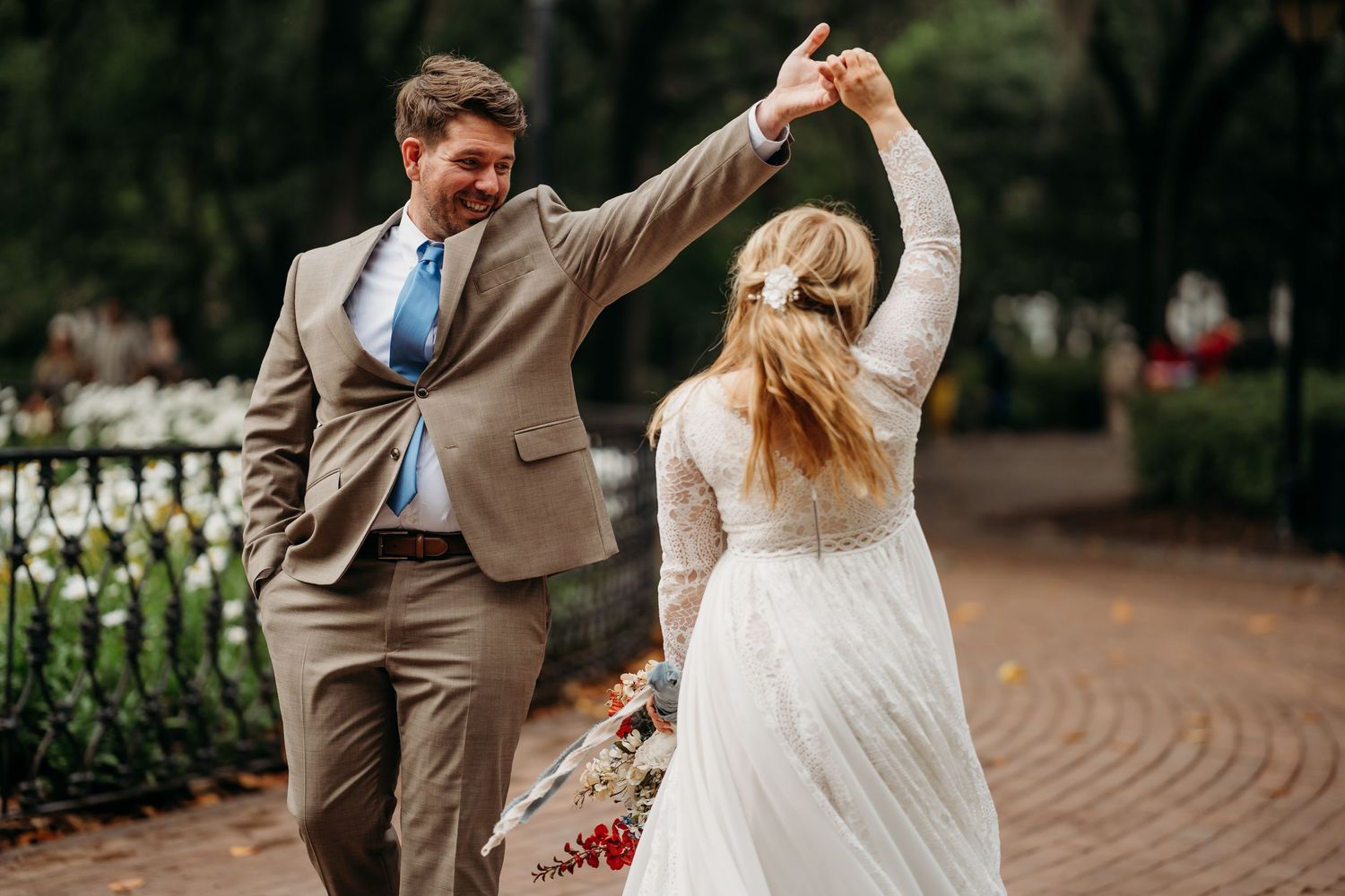 Smiling groom twirls his bride in a lace wedding gown on a tree-lined brick path surrounded by flowers.