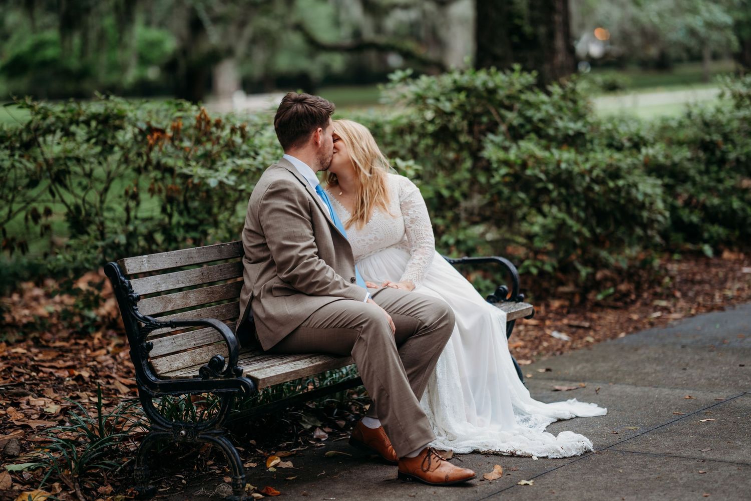Bride and groom sharing a tender kiss while seated on a park bench surrounded by autumn leaves.
