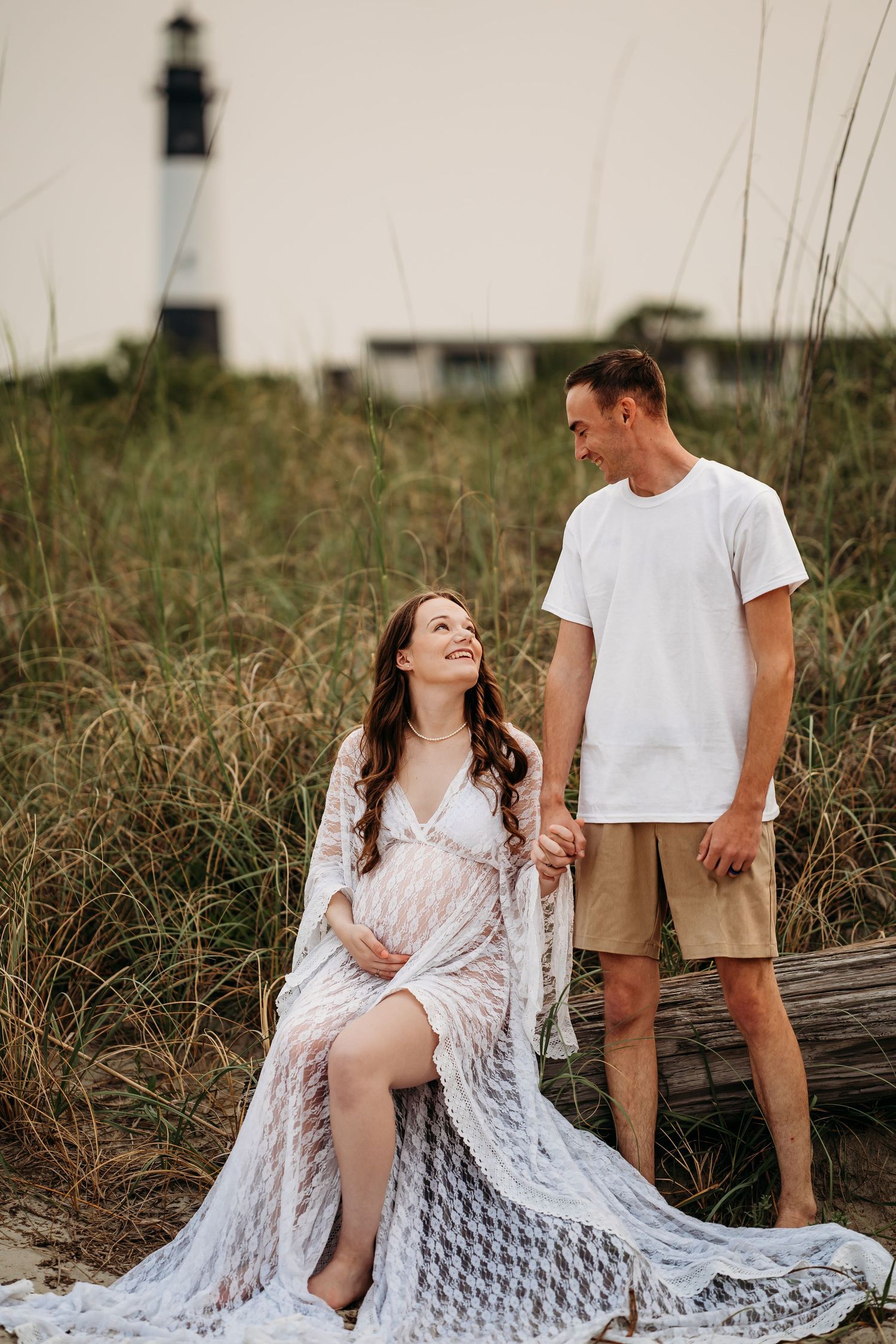 Pregnant woman in white lace gown sits on driftwood as partner stands beside her near lighthouse.