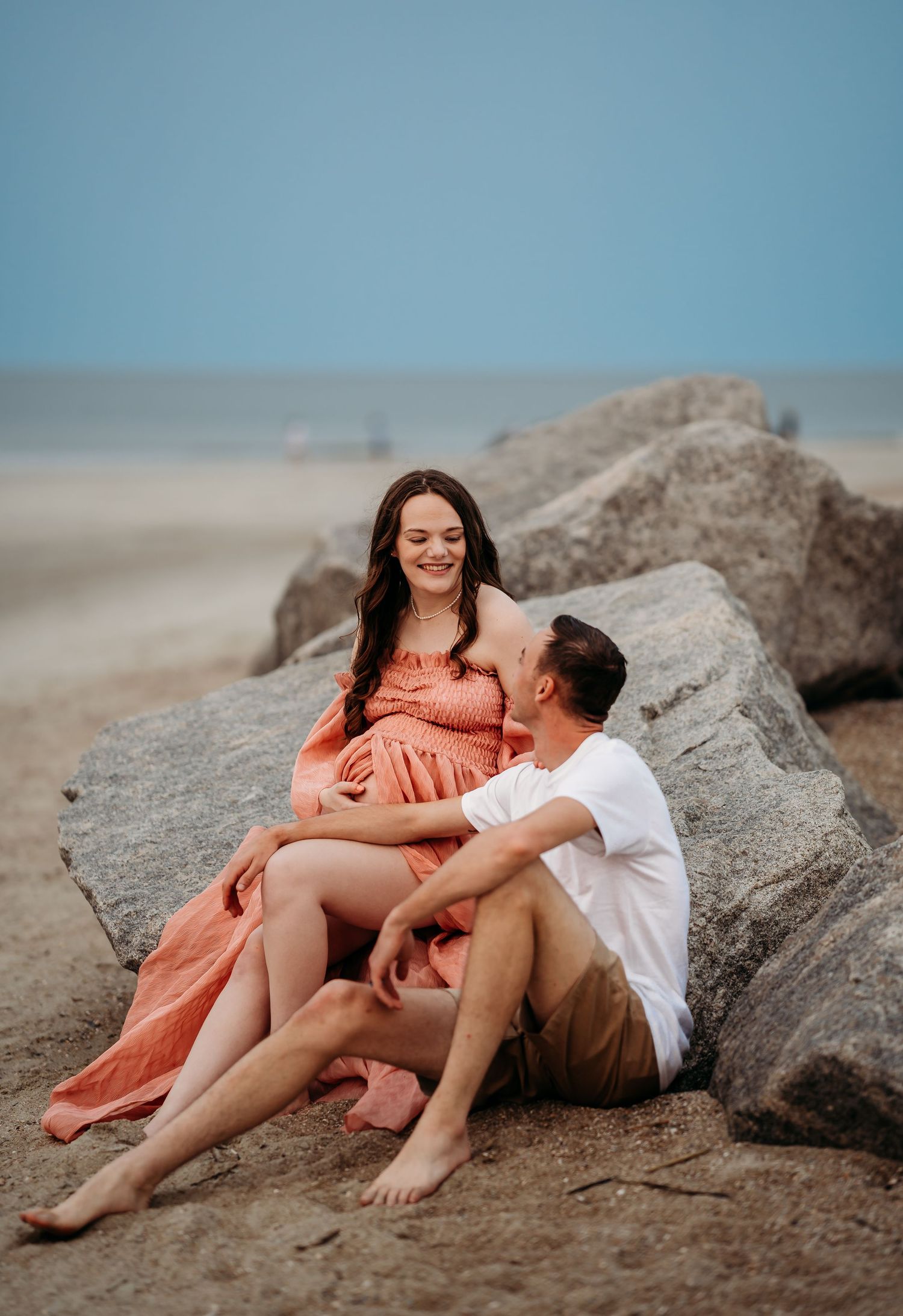 Pregnant woman in coral dress laughing with partner while relaxing on sandy beach rocks.