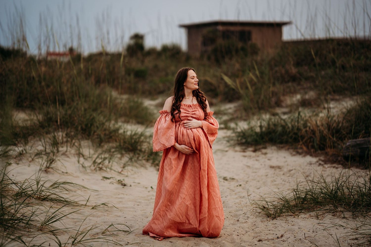 Pregnant woman in flowing coral maternity gown standing on sandy beach path surrounded by coastal dunes.
