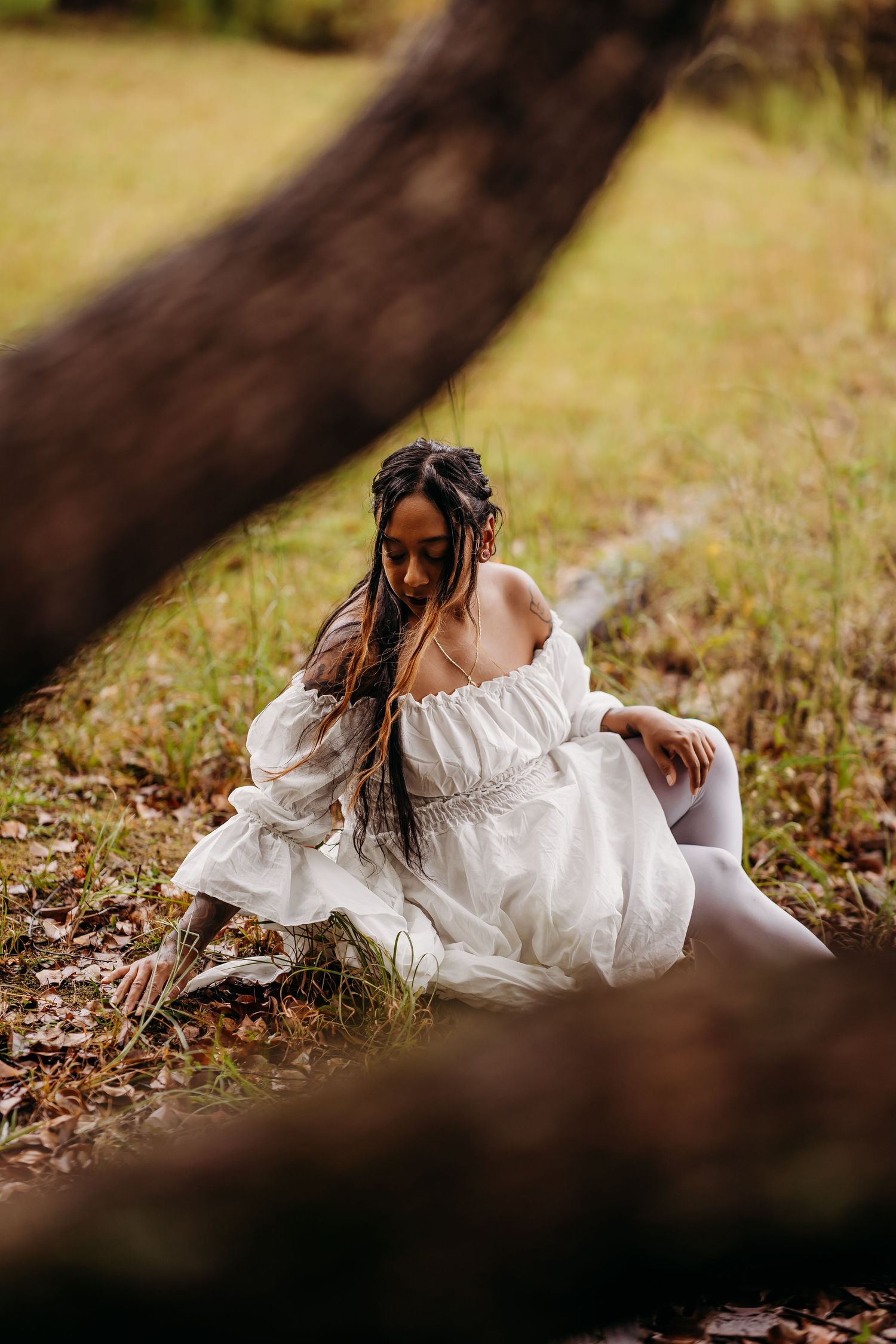 Woman in white off-shoulder dress sitting on the ground beneath a leaning tree outdoors.