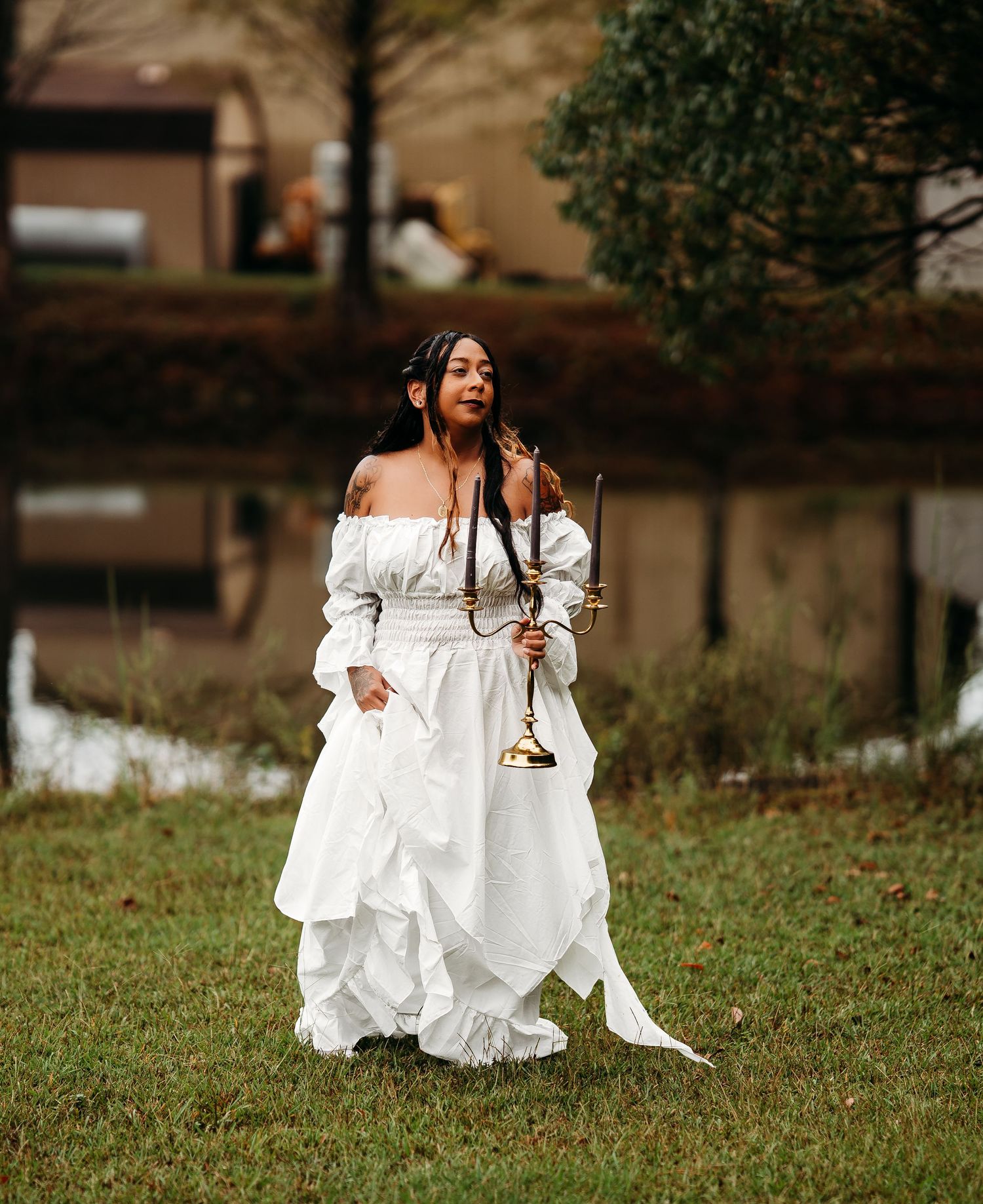 Woman in white off-shoulder dress stands on lawn holding gold candelabra with dark candles near building.