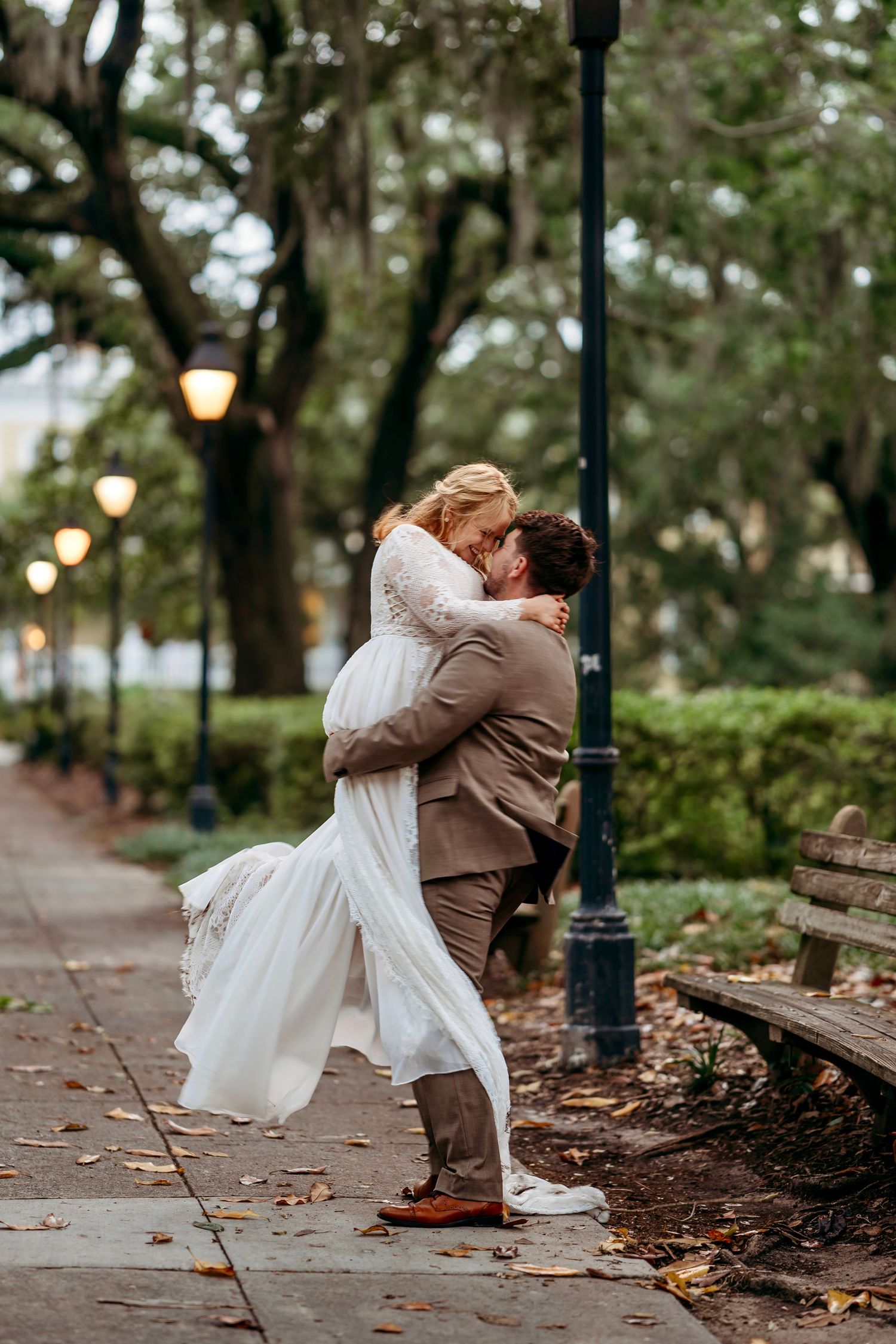 Groom lifting bride in white lace dress for a kiss in a park at dusk.