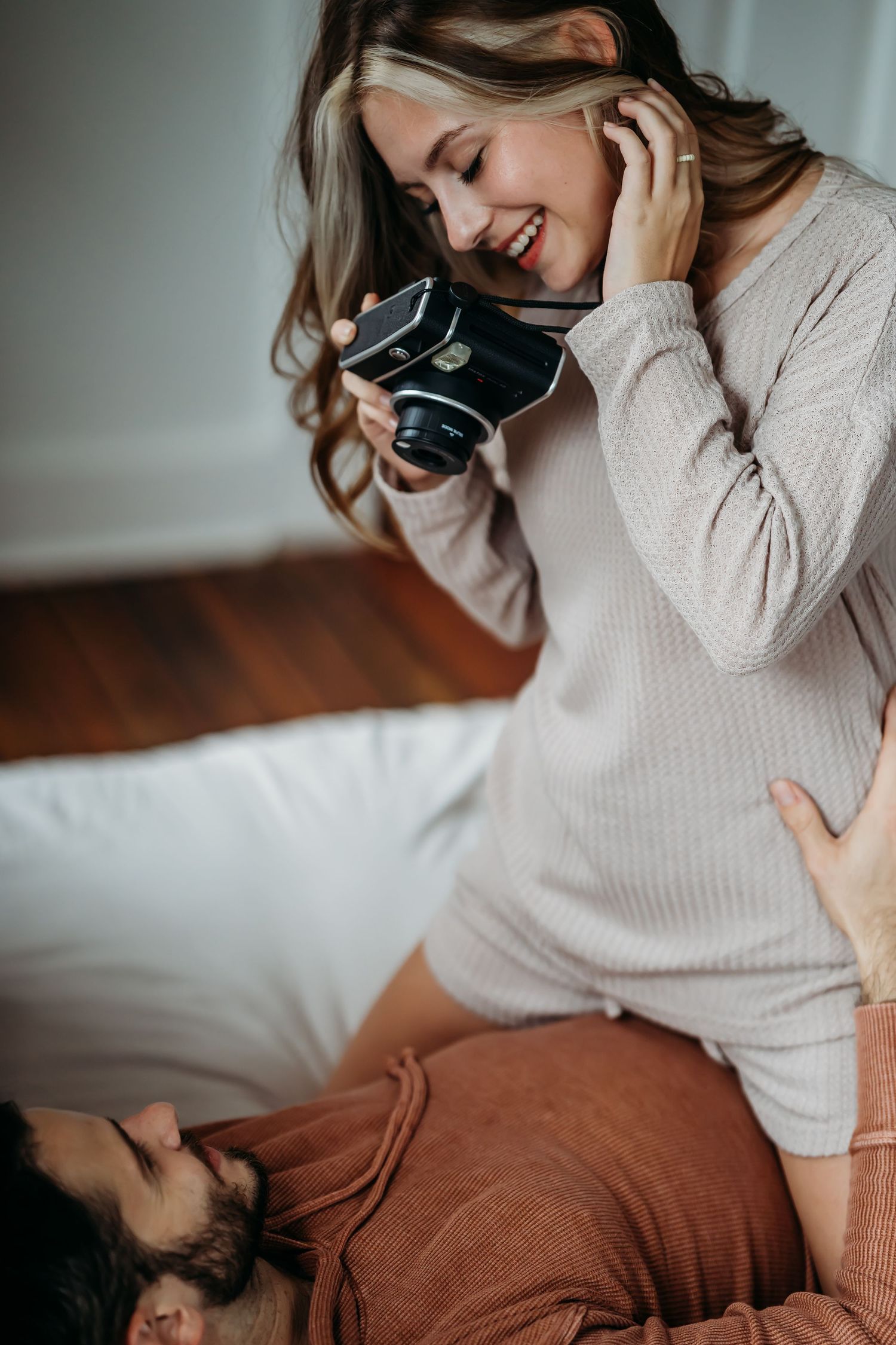 Smiling blonde woman photographing partner with vintage camera while sitting on bed.