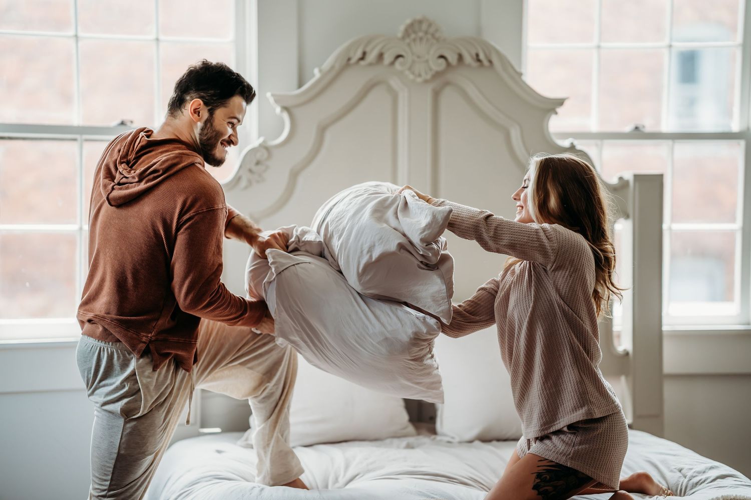 A joyful couple standing on a white bed engaged in a fun pillow fight near a sunlit window.