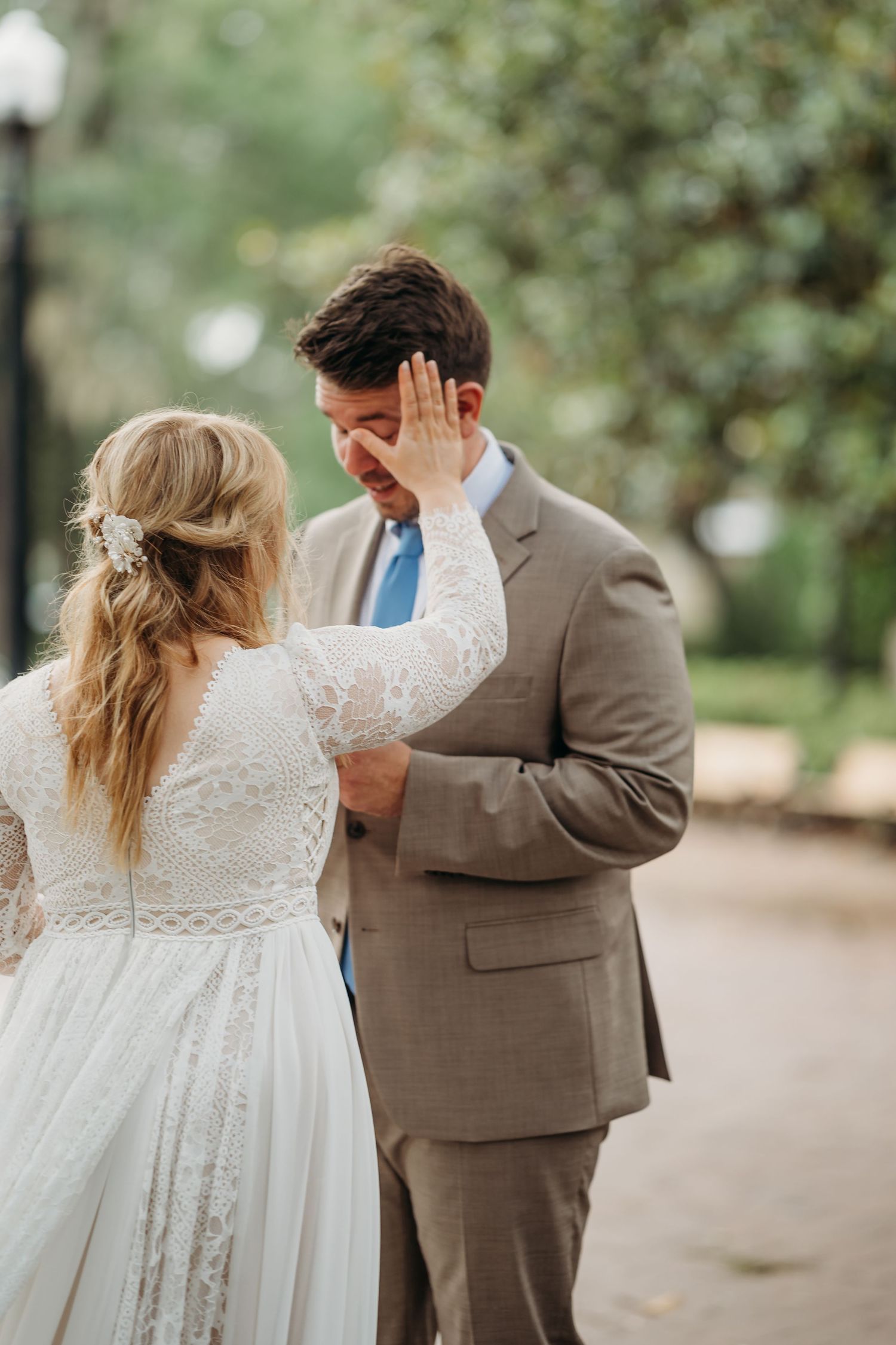 Bride in lace wedding gown tenderly wipes tears from emotional groom's face during outdoor ceremony.