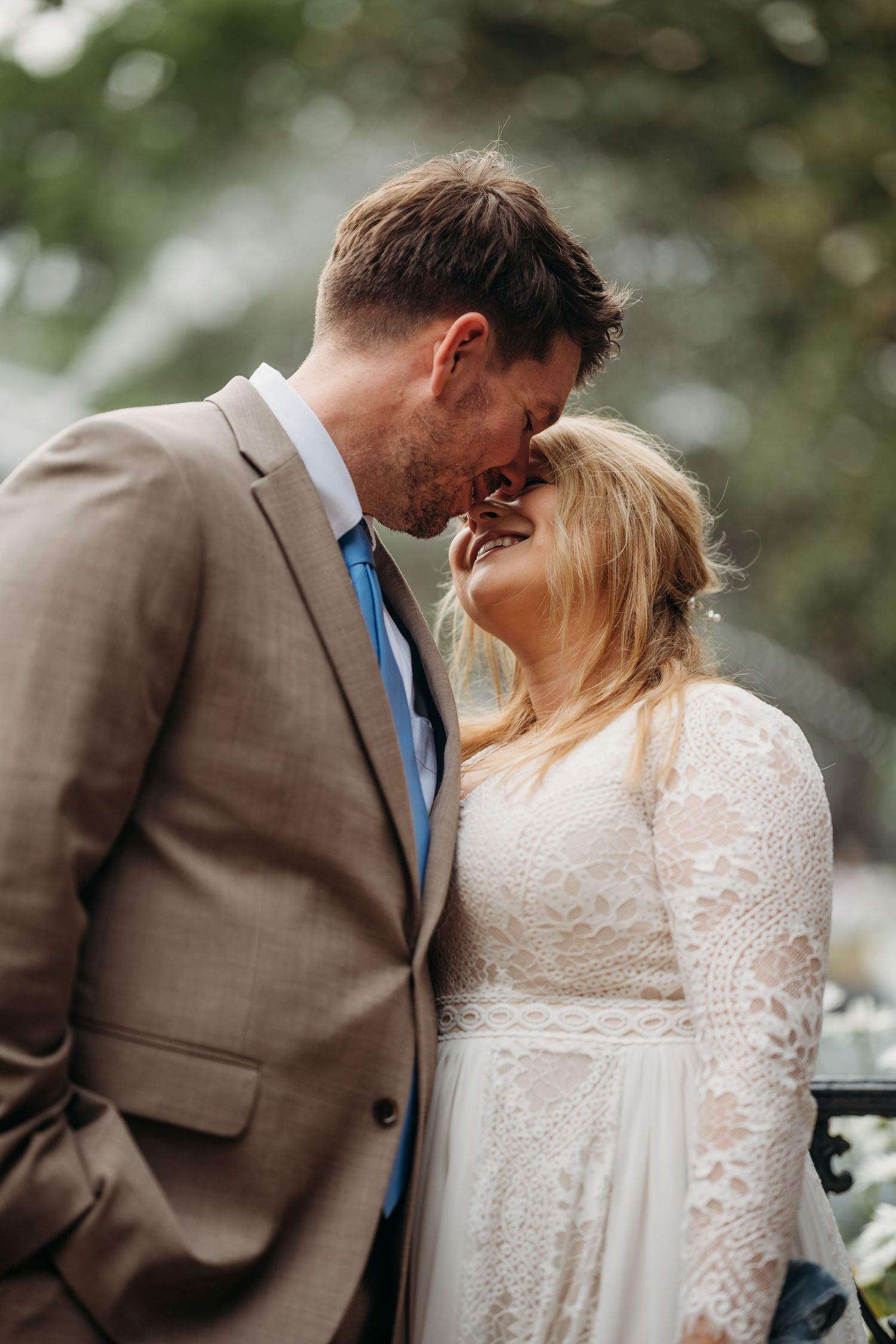 Smiling bride and groom lean foreheads together outdoors, groom in tan suit with blue tie, bride in long-sleeve lace gown.