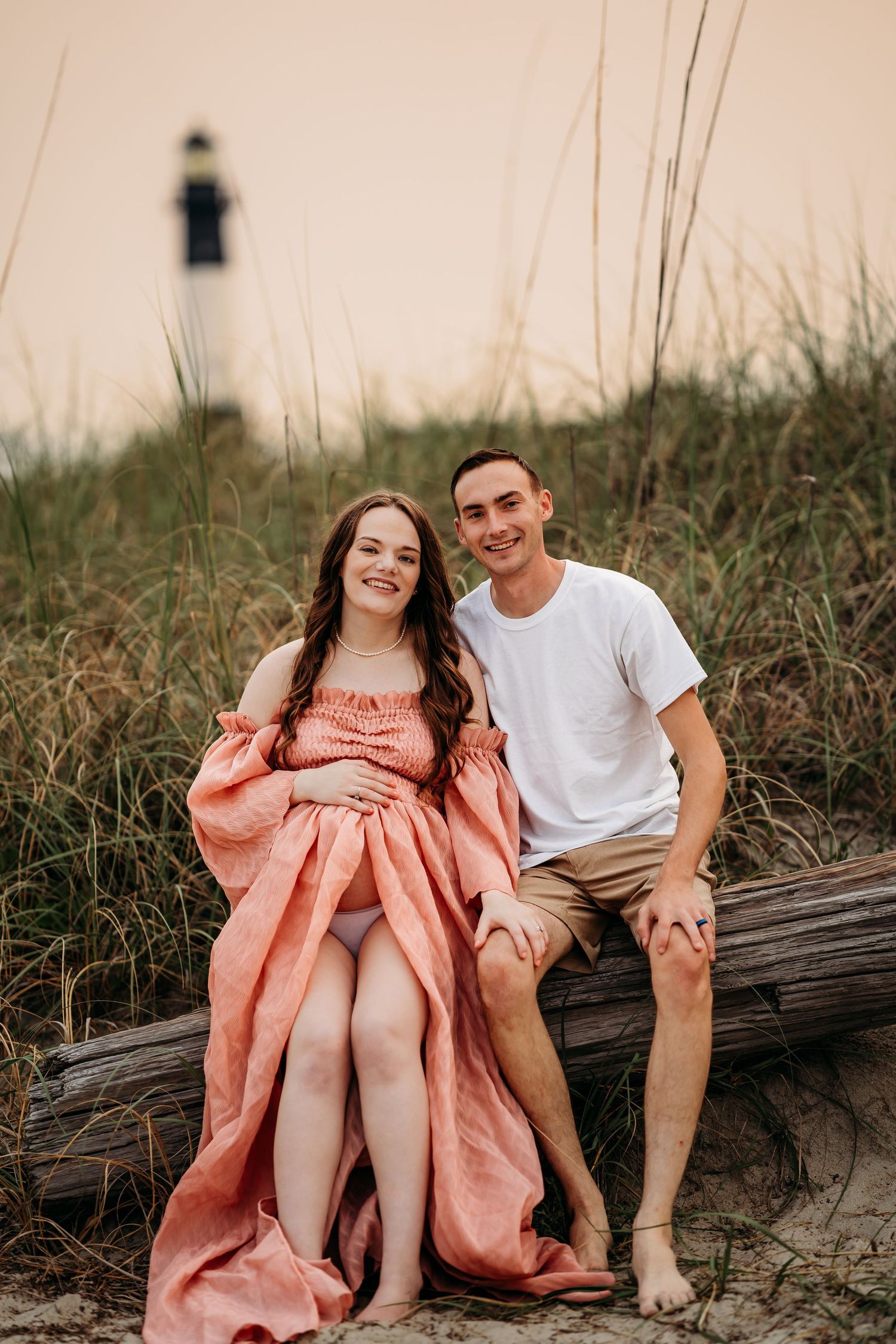 Smiling expecting couple sitting on driftwood with lighthouse backdrop during beach maternity shoot.